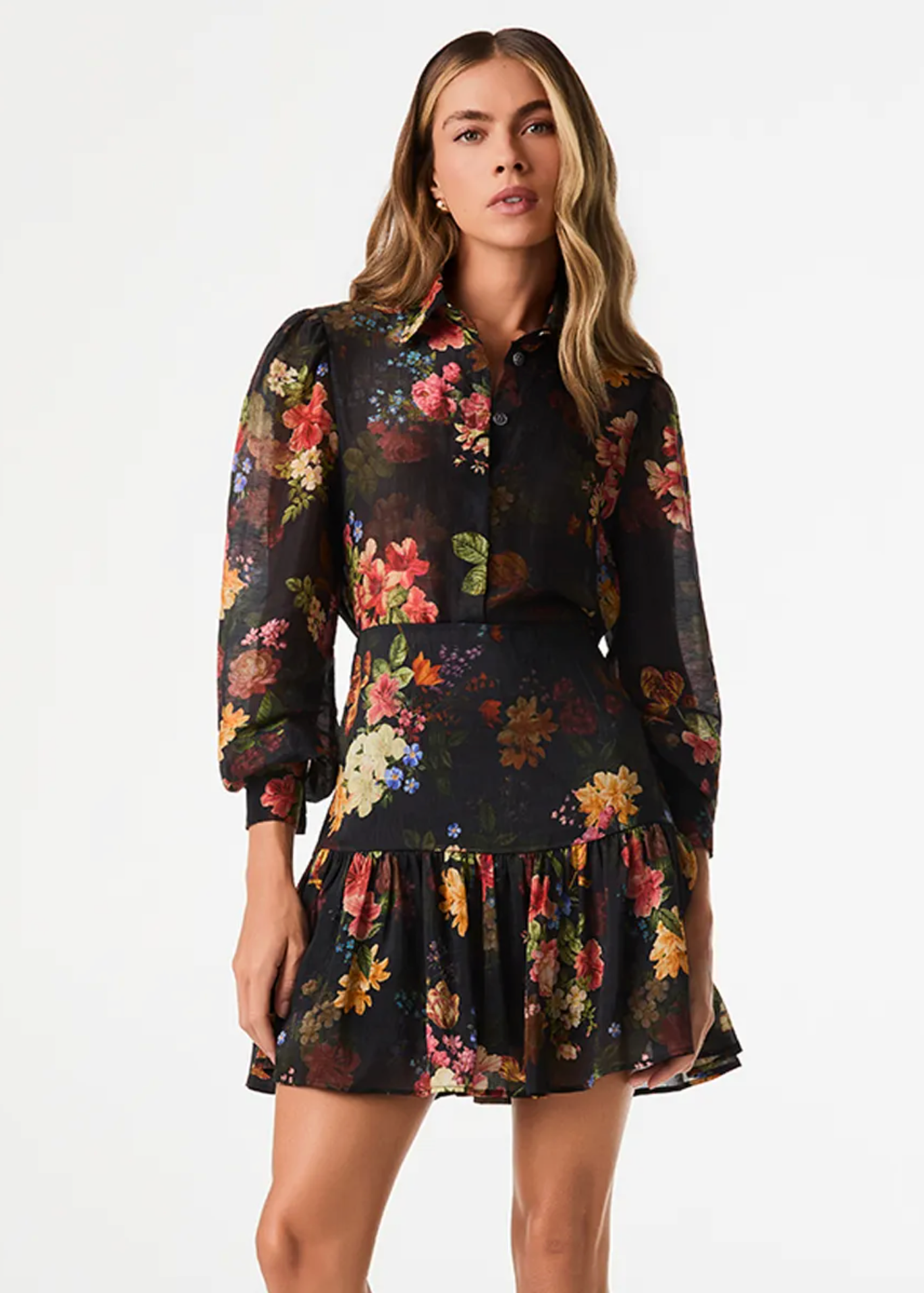 A woman modeling a black long-sleeve mini dress with a vibrant multicolor floral print, set against a plain white studio background.