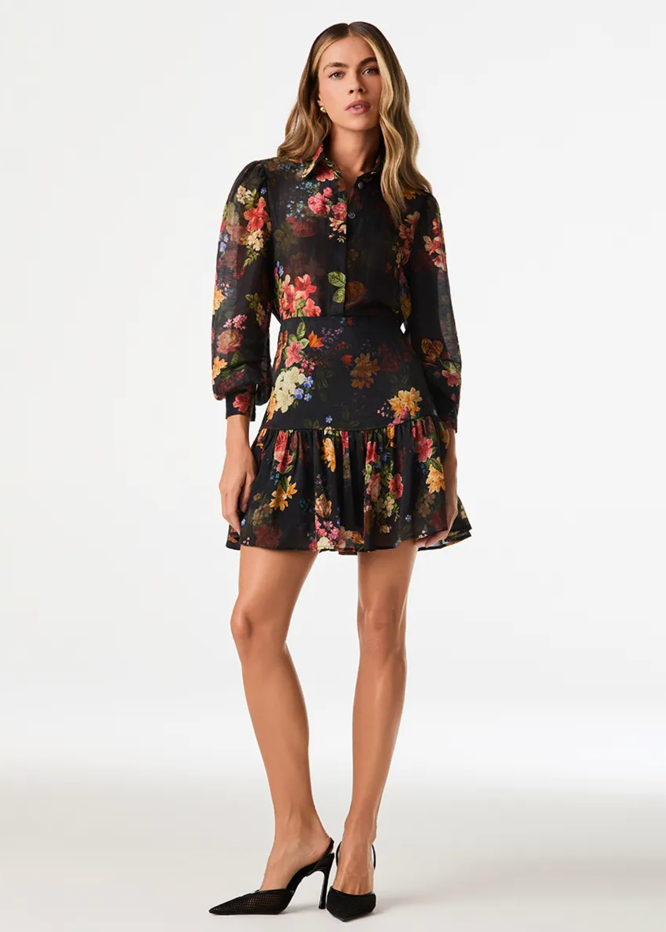A woman modeling a black long-sleeve mini dress with a vibrant multicolor floral print, paired with black pointed-toe heels, against a plain white studio background.