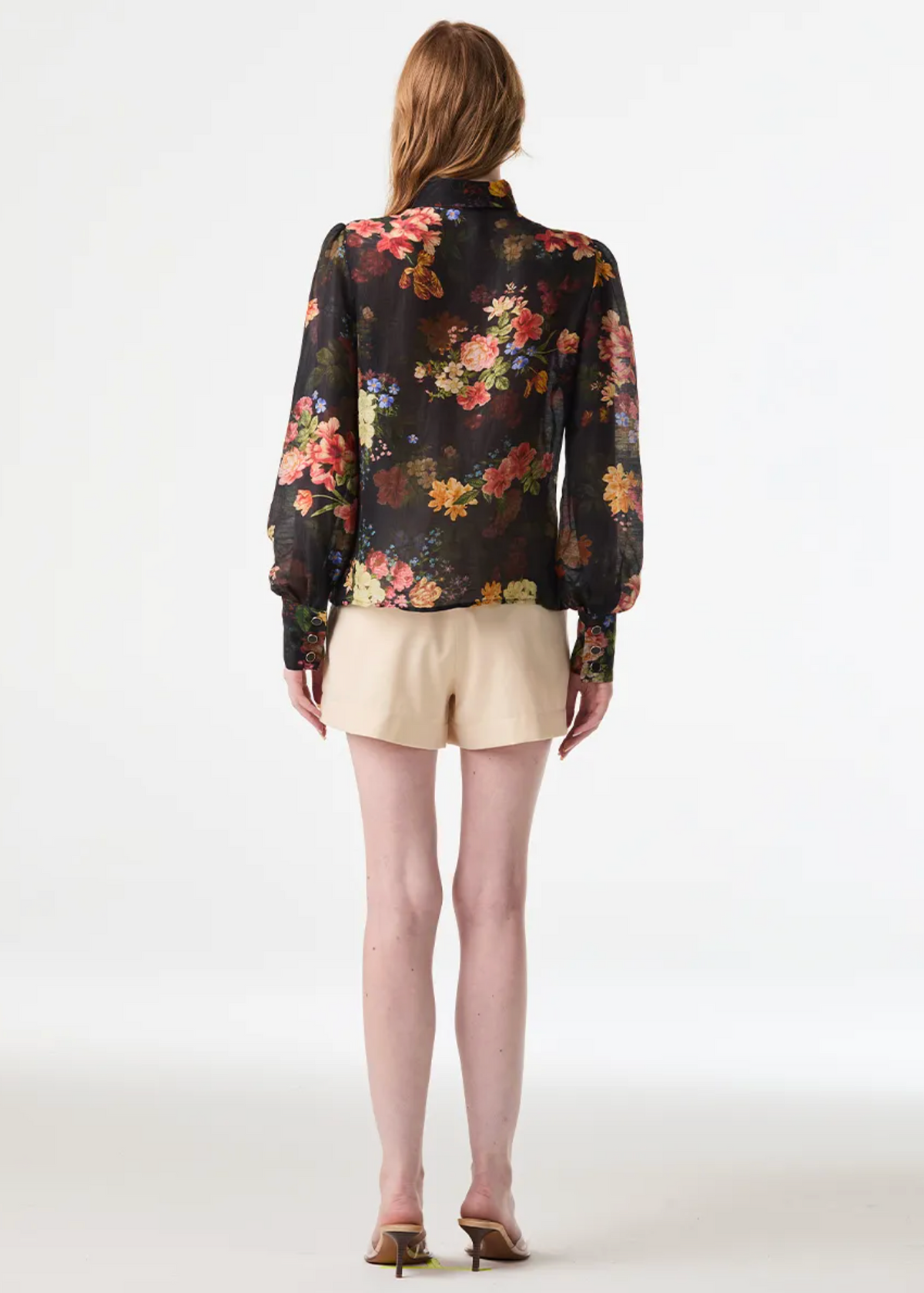 A woman is shown from the back wearing a black long-sleeve blouse with a vibrant multicolor floral print, paired with beige shorts and brown heels, against a plain white studio background.