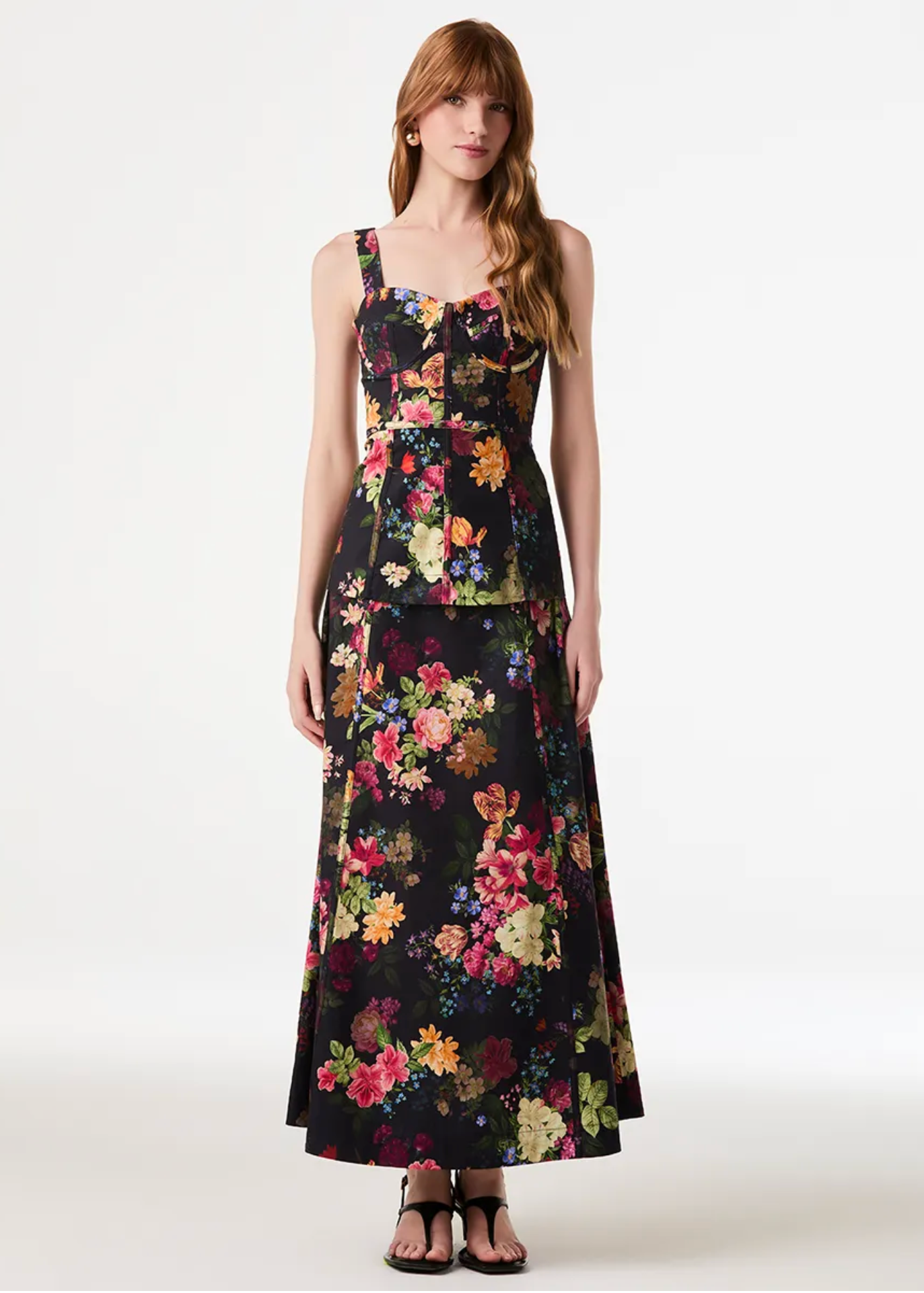 A woman modeling a sleeveless black maxi dress with a vibrant multicolor floral print, photographed against a seamless white studio background.