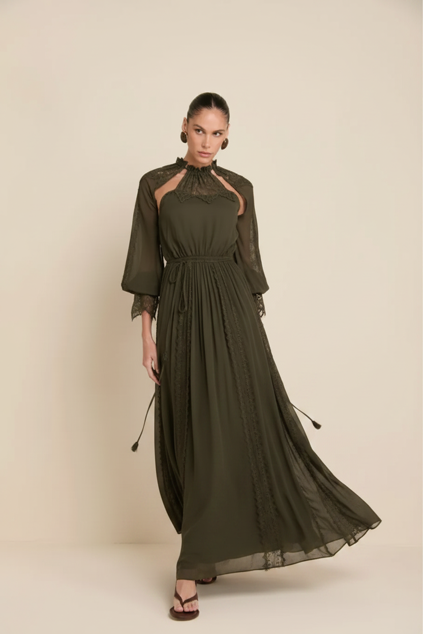 A woman modeling a long, dark green midi dress with sheer lace details on the shoulders and sleeves, standing against a solid beige studio background.