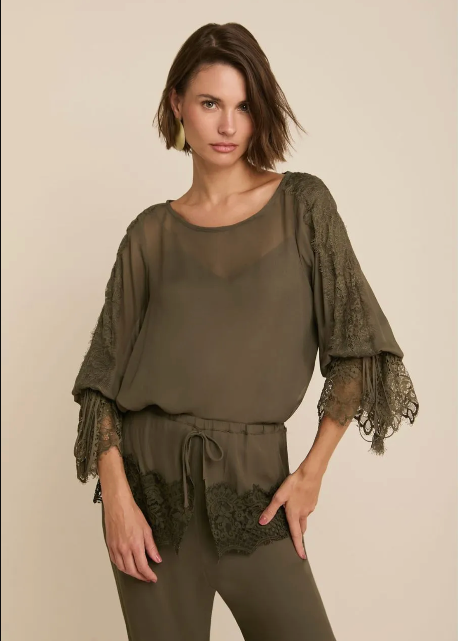 A woman modeling an olive green two-piece set featuring a sheer long-sleeve top with lace flutter sleeves and matching drawstring pants, set against a solid beige background.