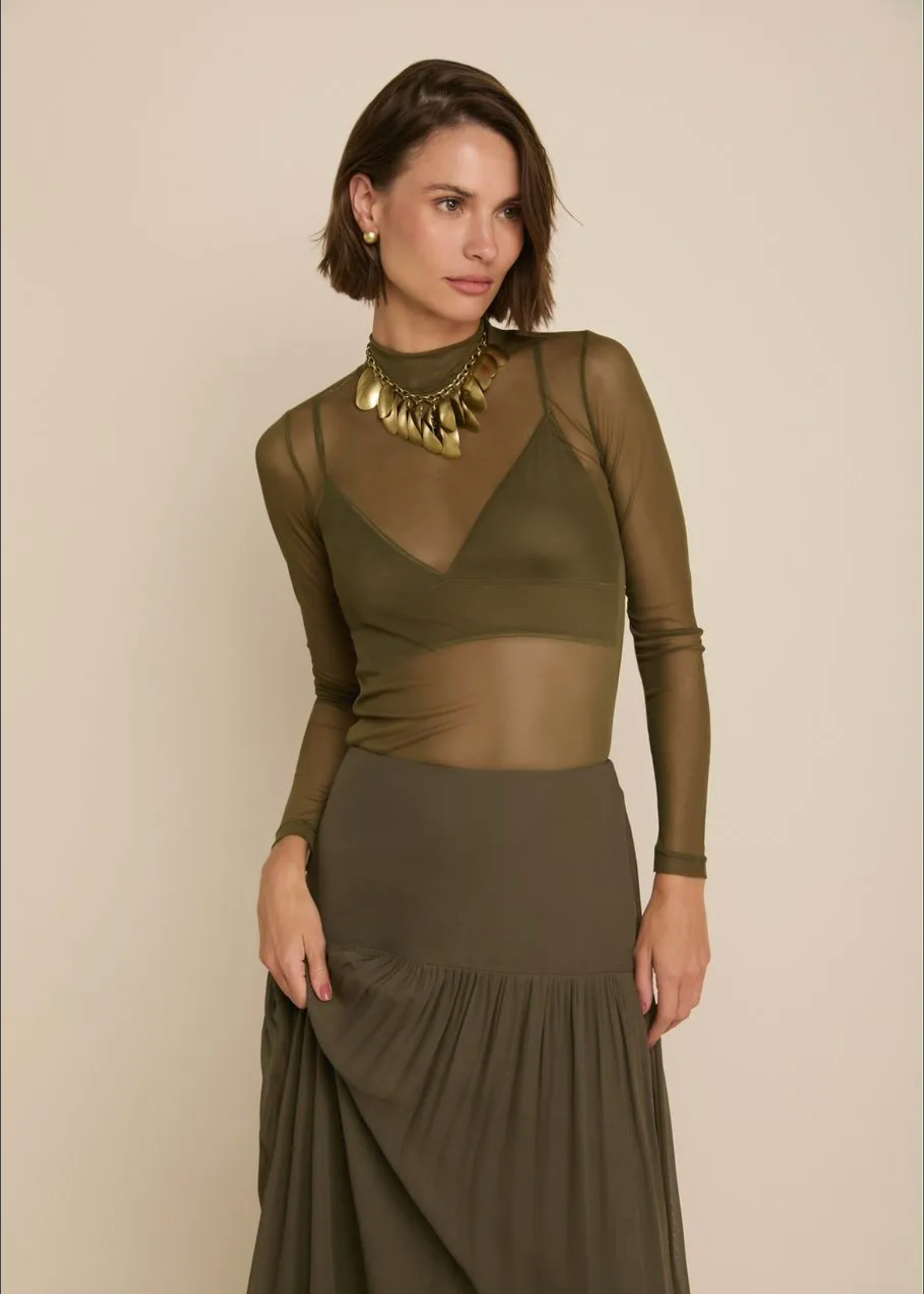 A woman modeling an olive green long-sleeve mesh top with a matching pleated maxi skirt, styled with a chunky gold statement necklace against a solid beige background.