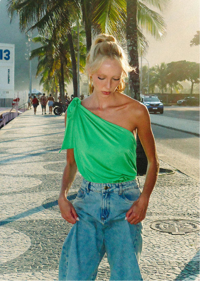 Woman wearing a green top and blue jeans on a beach with palm trees and people in the background.