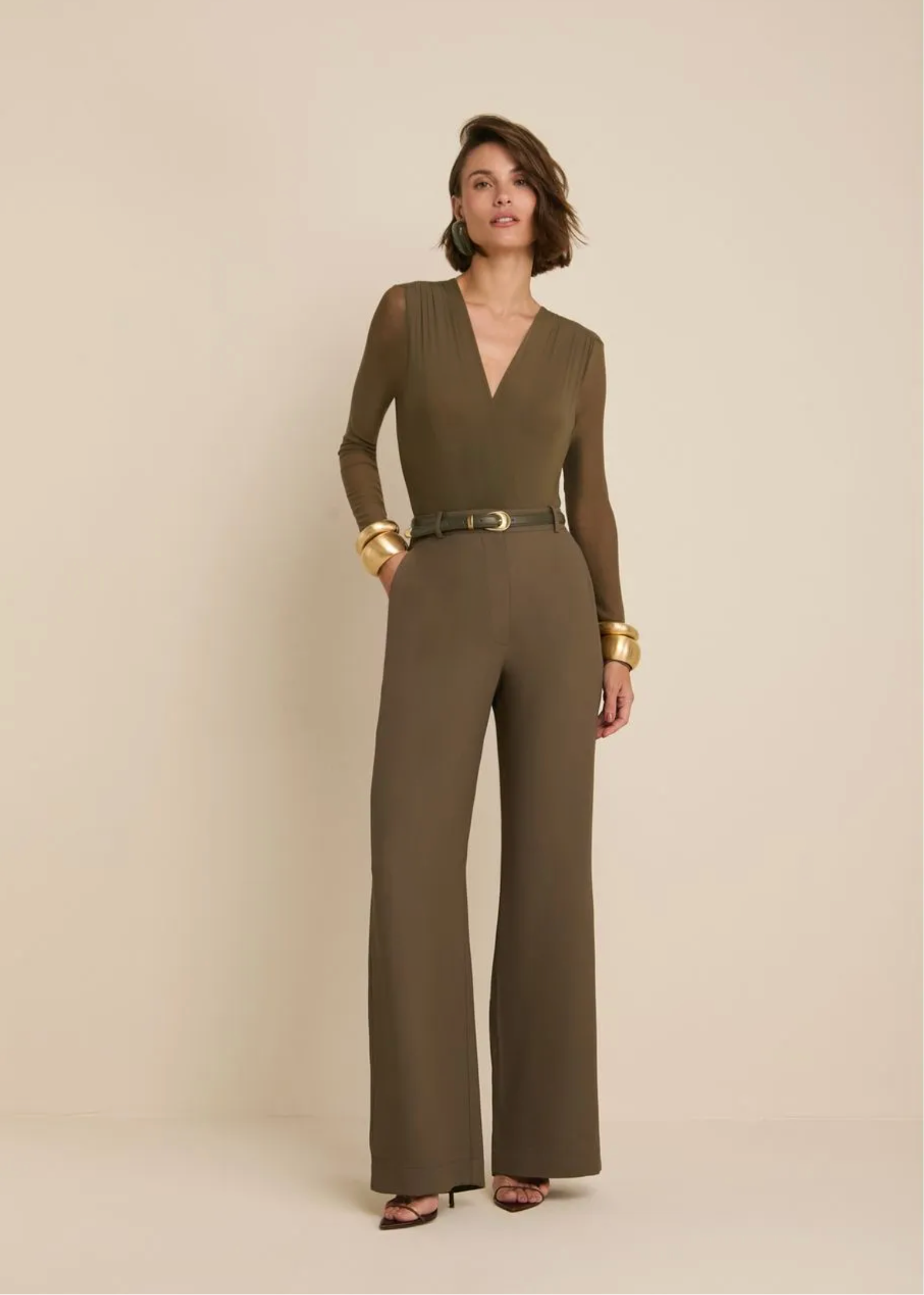 A woman modeling a long-sleeve olive brown wide-leg jumpsuit with a matching belt, styled with gold bracelets, against a solid beige studio background.