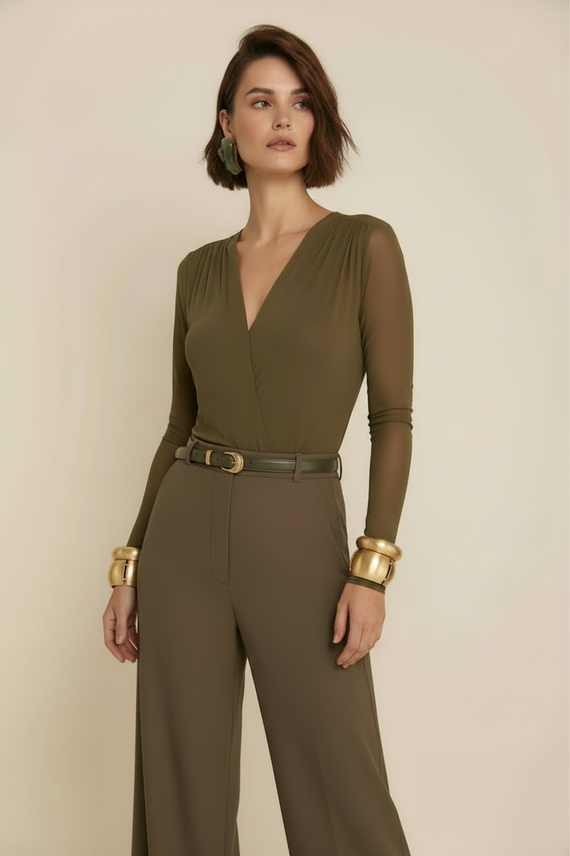 A woman modeling a solid olive green long-sleeve jumpsuit with a matching belt and gold-tone cuffs, set against a plain beige studio background.