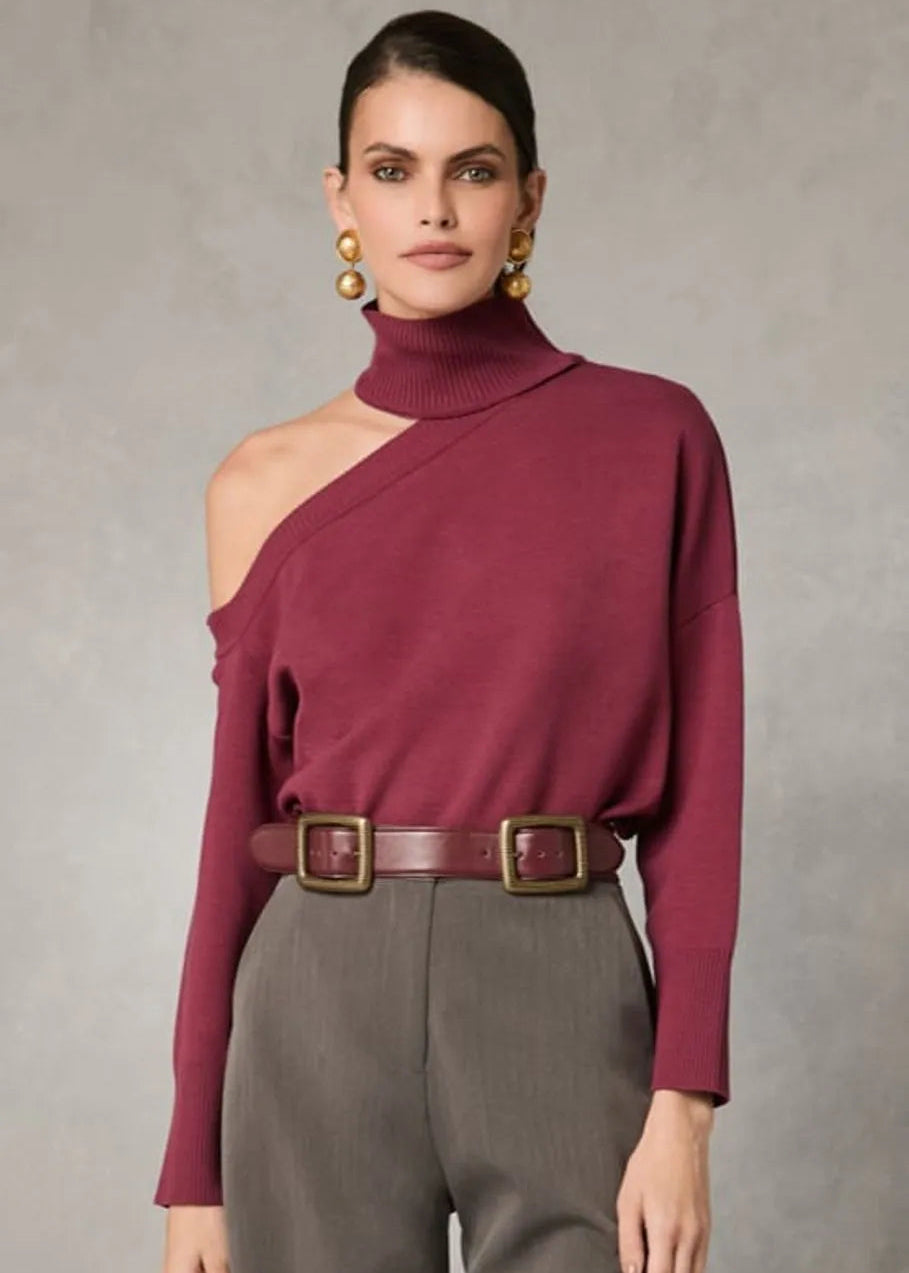 Woman wearing a burgundy sweater with one shoulder and gray pants against a neutral background