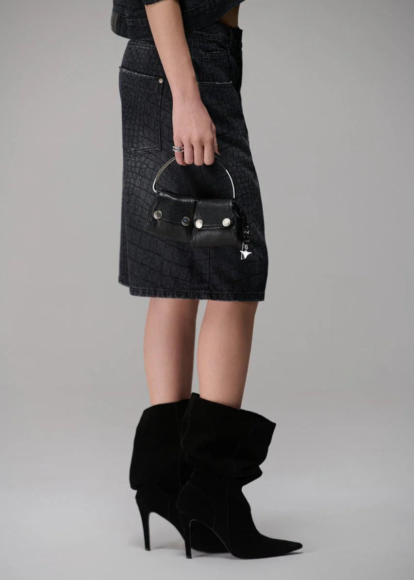 Person wearing a black denim skirt and black high-heeled boots, holding a small black handbag.