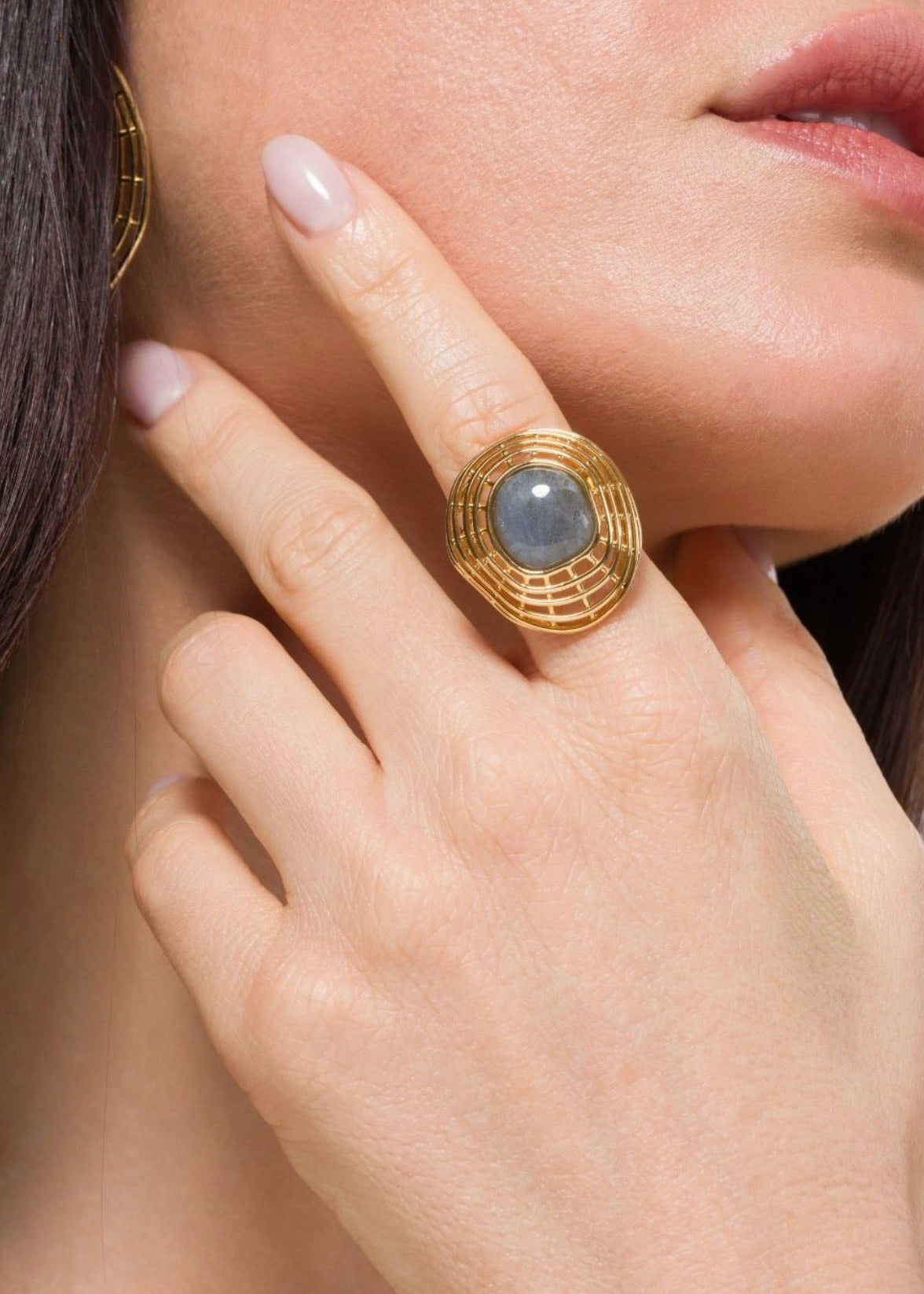 A close-up lifestyle shot of a model wearing a large gold ring with a round gray-blue stone, with matching gold hoop earrings partially visible.