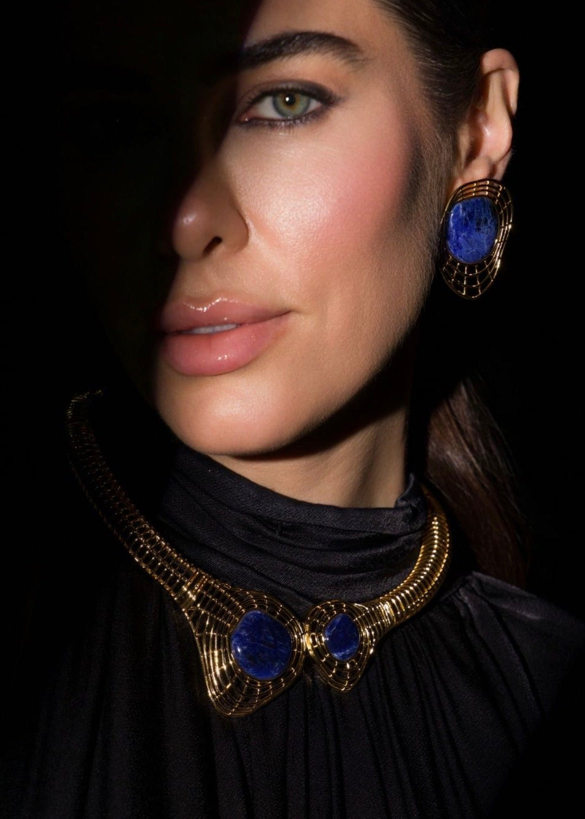 A close-up portrait of a woman wearing a gold-toned statement necklace and matching drop earrings with deep blue cabochon stones, set against a dark background.