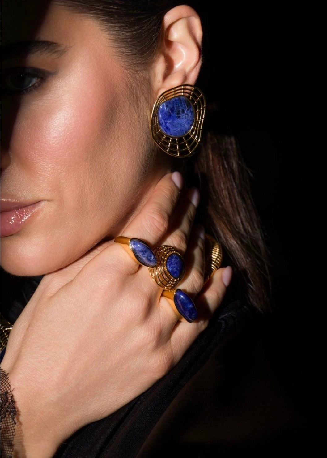 A close-up lifestyle shot of a woman wearing gold jewelry, featuring a large gold hoop earring with a round blue lapis lazuli stone and matching gold rings with oval blue stones on her fingers.
