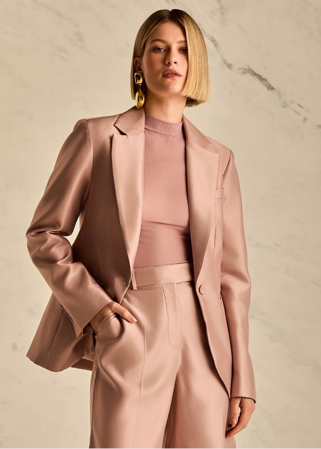 A woman modeling a matching blush pink satin three-piece suit, featuring a structured blazer, a fitted mock-neck top, and tailored trousers, set against a light beige marble wall.