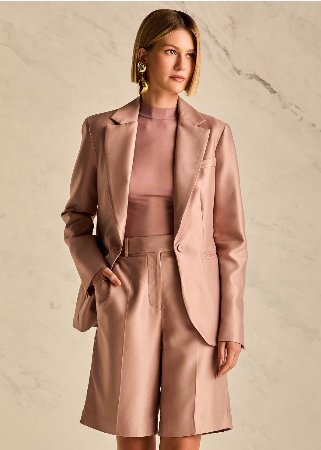 A woman models a matching dusty rose satin three-piece suit consisting of a blazer, high-neck top, and wide-leg shorts, set against a light beige marble background.