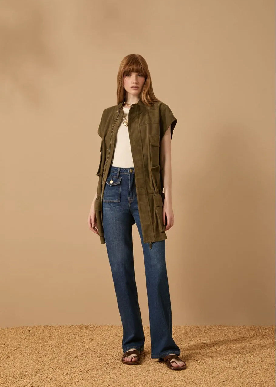 Woman wearing a green vest over a white shirt and blue jeans on a beige background