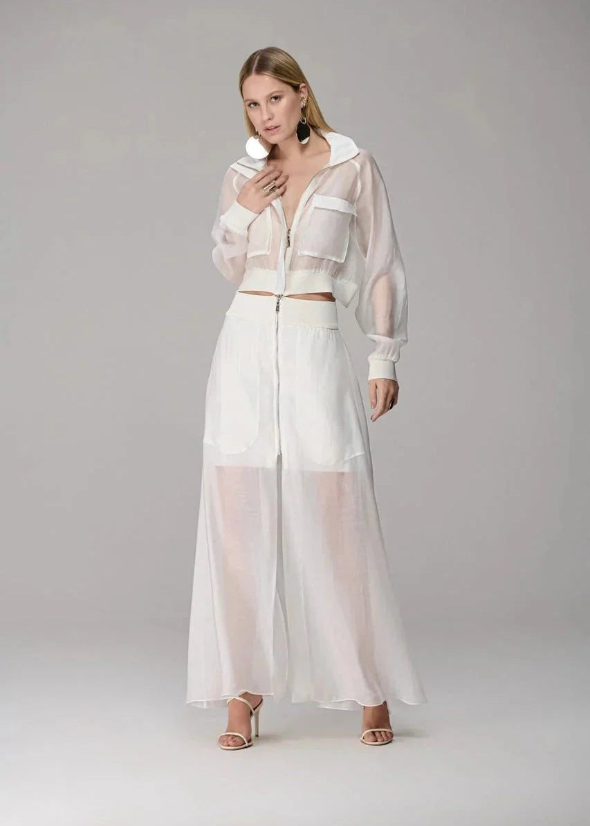 A model wearing a white two-piece outfit consisting of a sheer long-sleeve cropped zip-up jacket and matching wide-leg sheer maxi pants, photographed against a neutral light gray studio background.