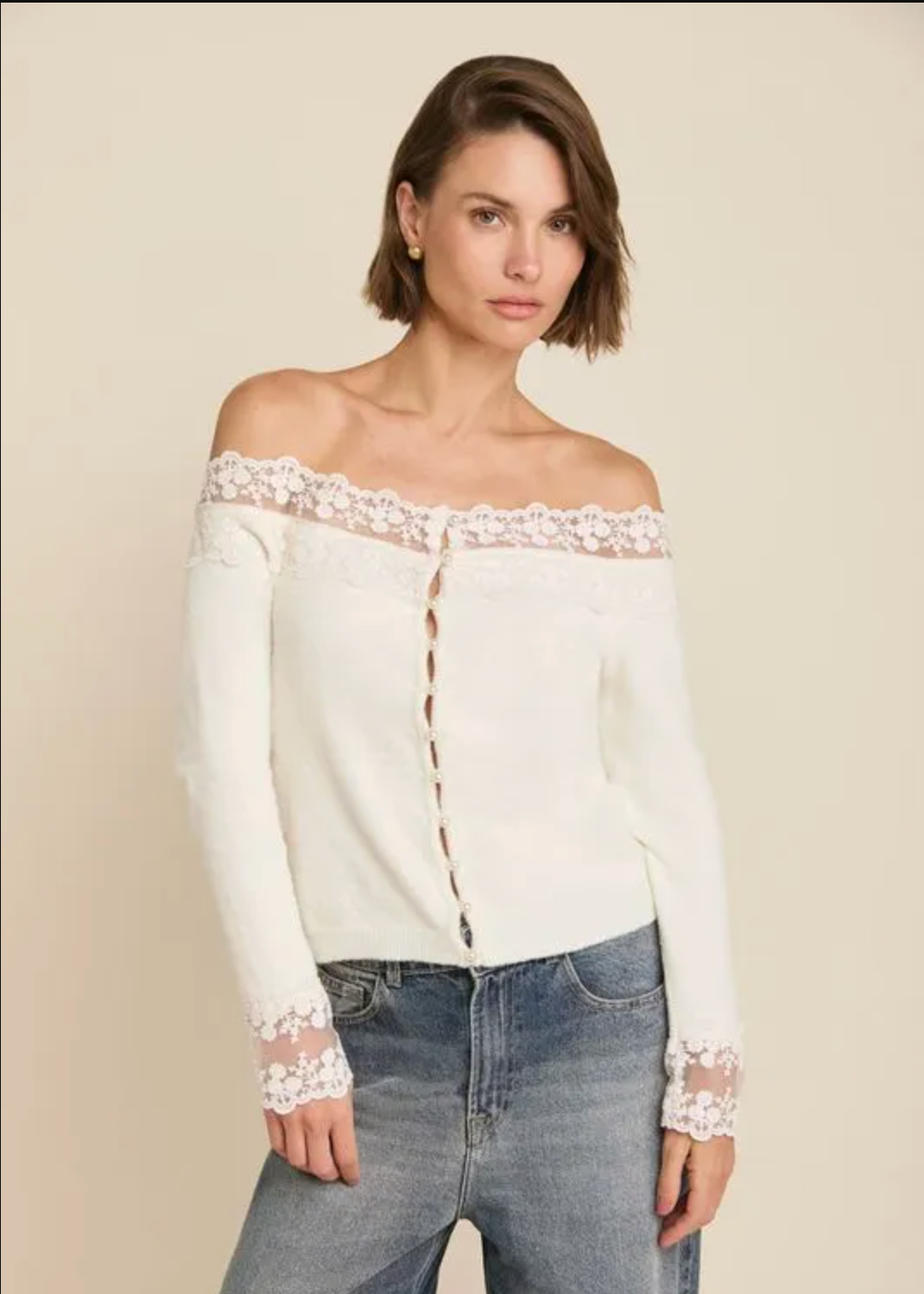 A woman modeling an off-the-shoulder cream knit top with delicate lace trim along the neckline and cuffs, paired with blue jeans, against a solid light beige background.