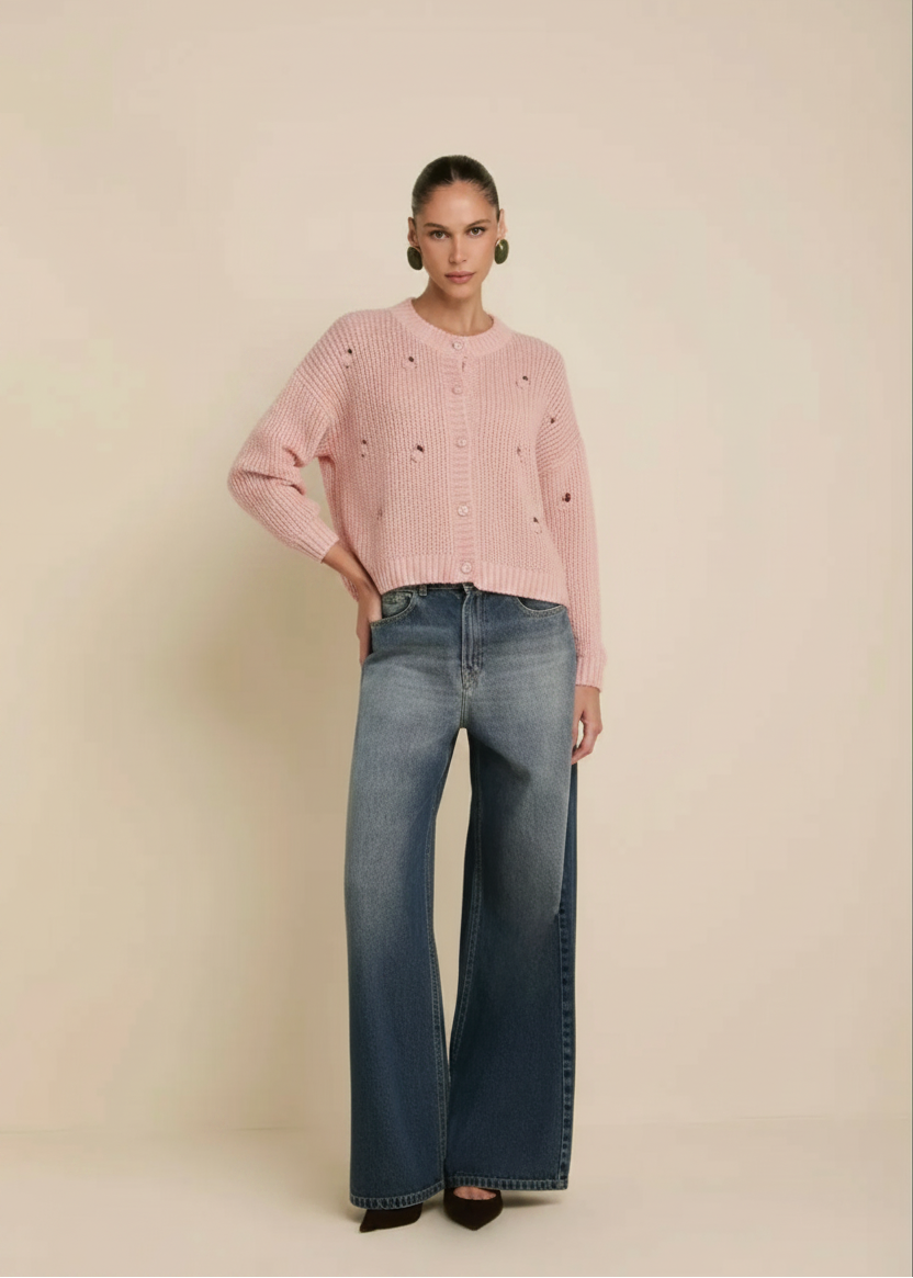 A model stands against a plain beige studio background wearing a cream-colored textured knit sweater with small black embroidered details, paired with wide-leg blue jeans.