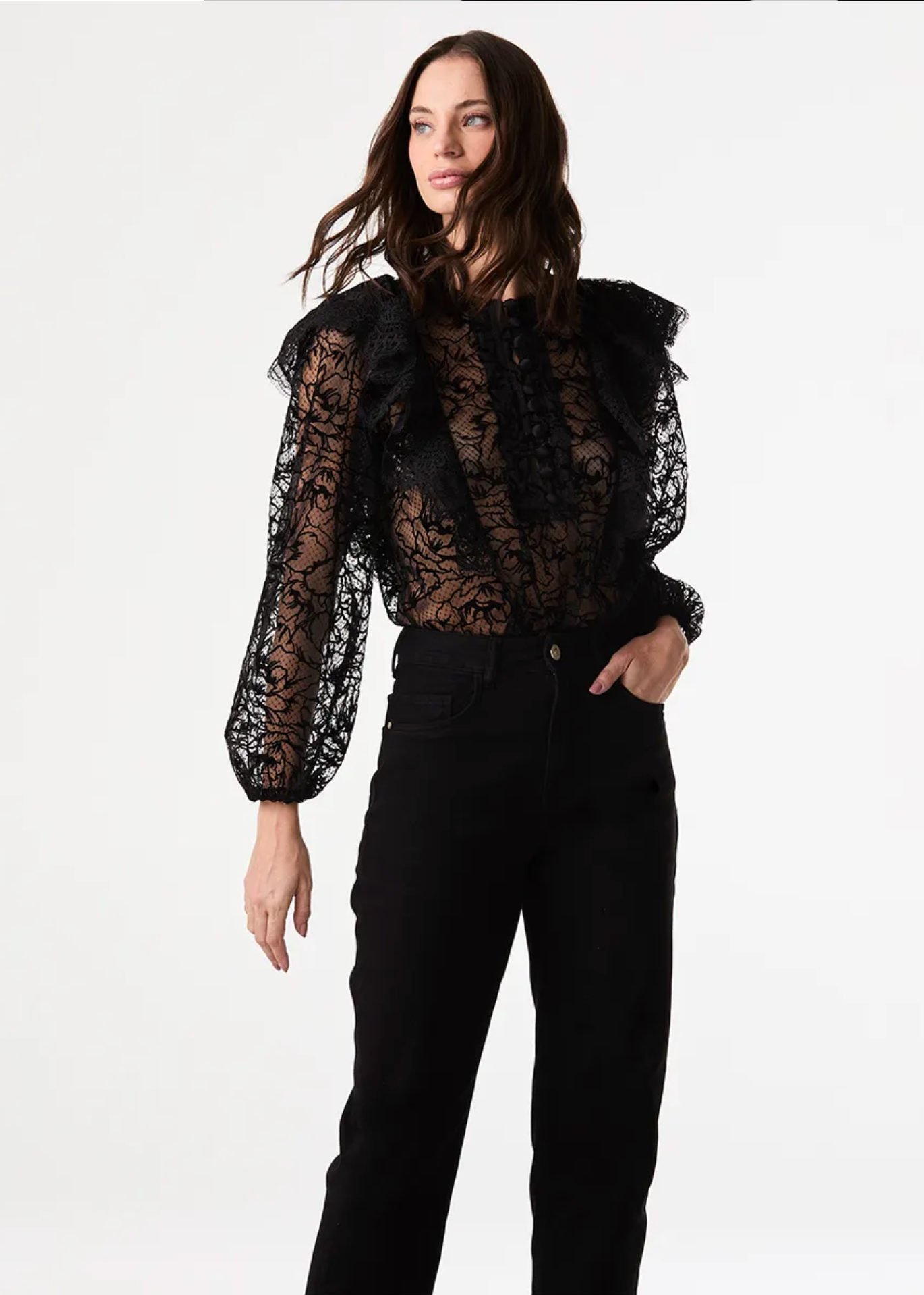 A woman modeling a black sheer lace long-sleeve blouse with dramatic ruffled shoulders and black pants, photographed against a clean white studio background.
