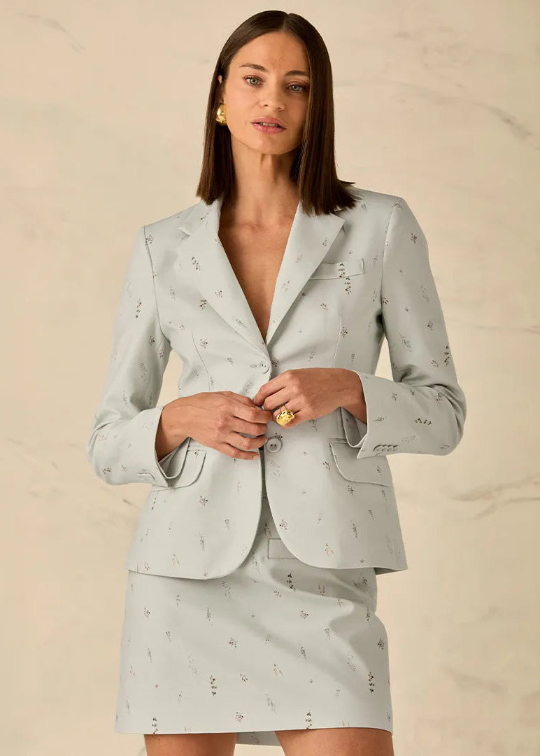 A woman models a light sage green two-piece suit with a small black floral pattern, featuring a blazer and matching mini skirt, set against a neutral beige marble background.