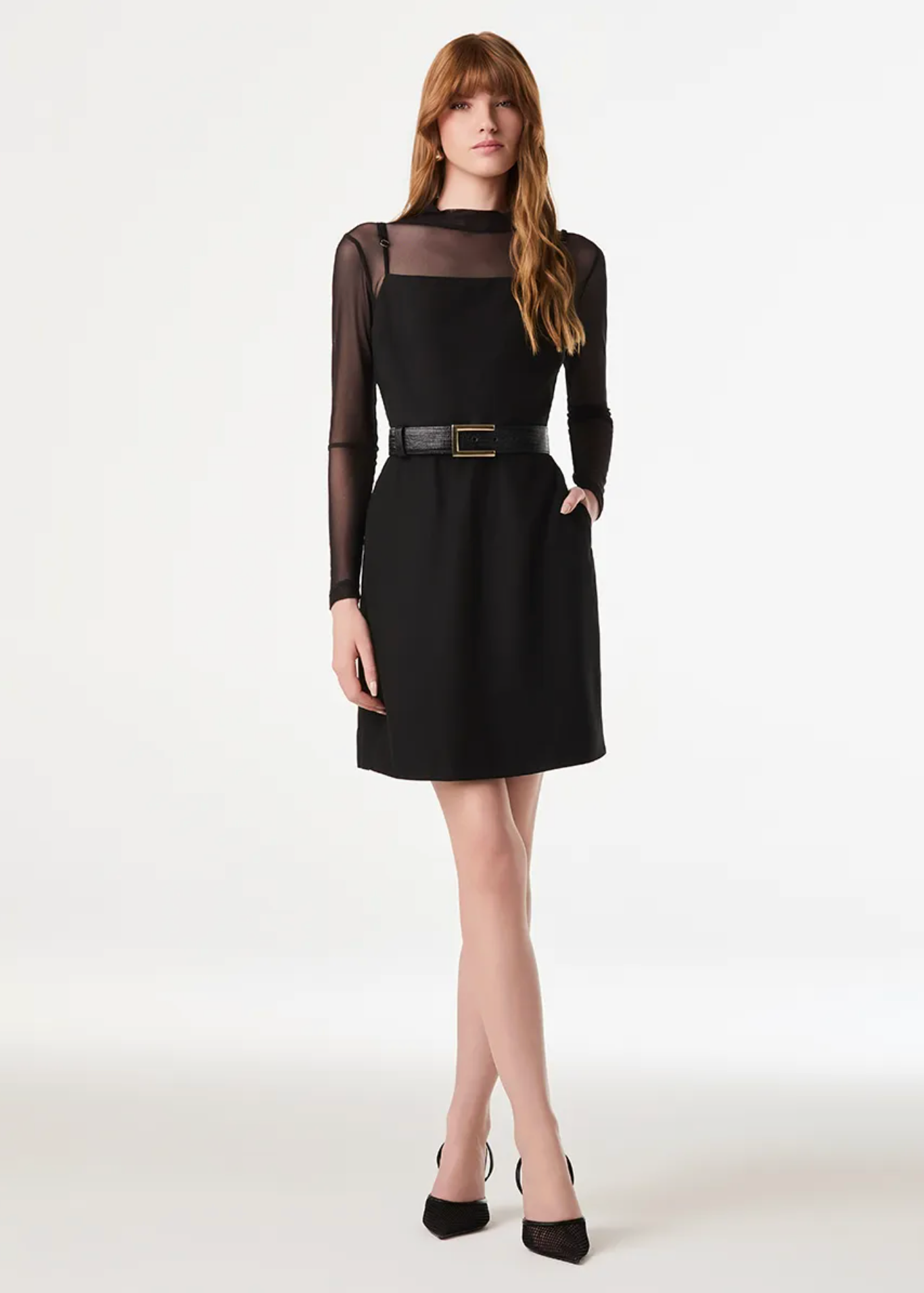A woman modeling a black long-sleeve shift dress with a sheer mesh yoke and a thin black belt with a gold buckle, paired with black heels, against a plain white studio background.