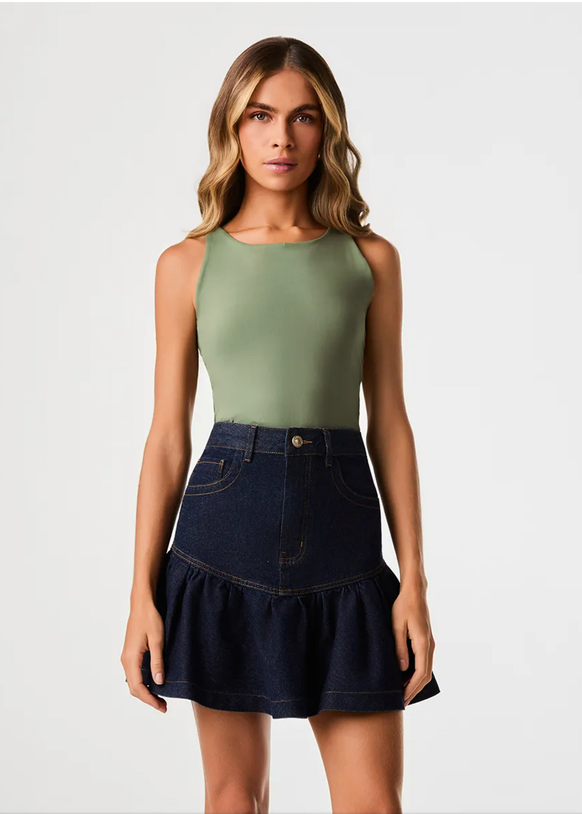 A woman modeling a sleeveless sage green top paired with a dark blue denim mini skirt with a ruffled hem, shot against a plain light gray studio background.