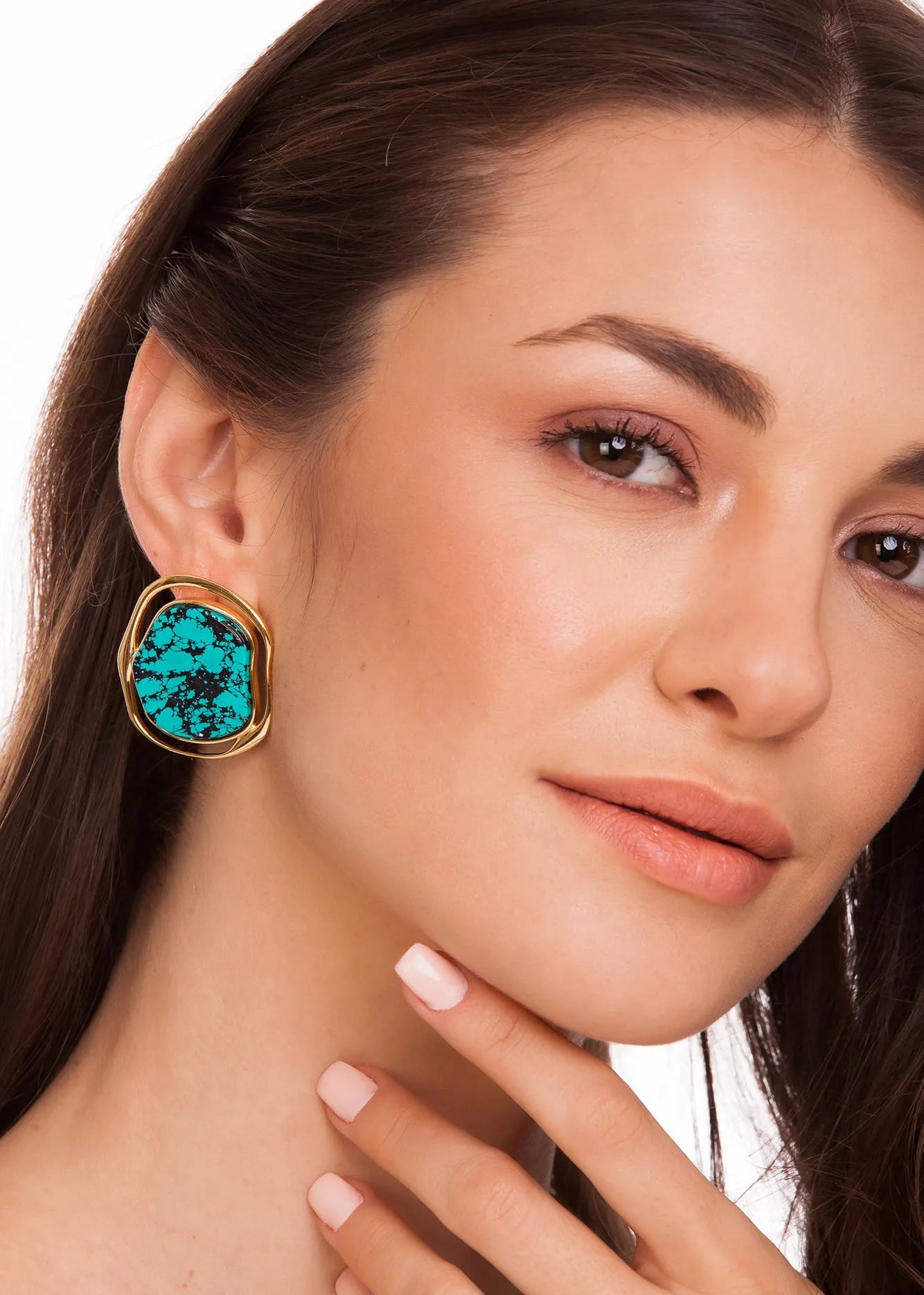 A close-up of a woman wearing a large gold-tone statement earring with a vibrant turquoise blue-green stone, set against a clean white background.