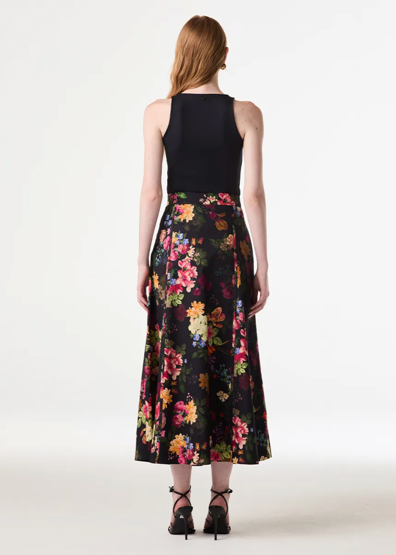 A woman shown from the back wearing a sleeveless black top and a high-waisted black midi skirt with a colorful floral print, paired with black strappy heels, against a plain white studio background.