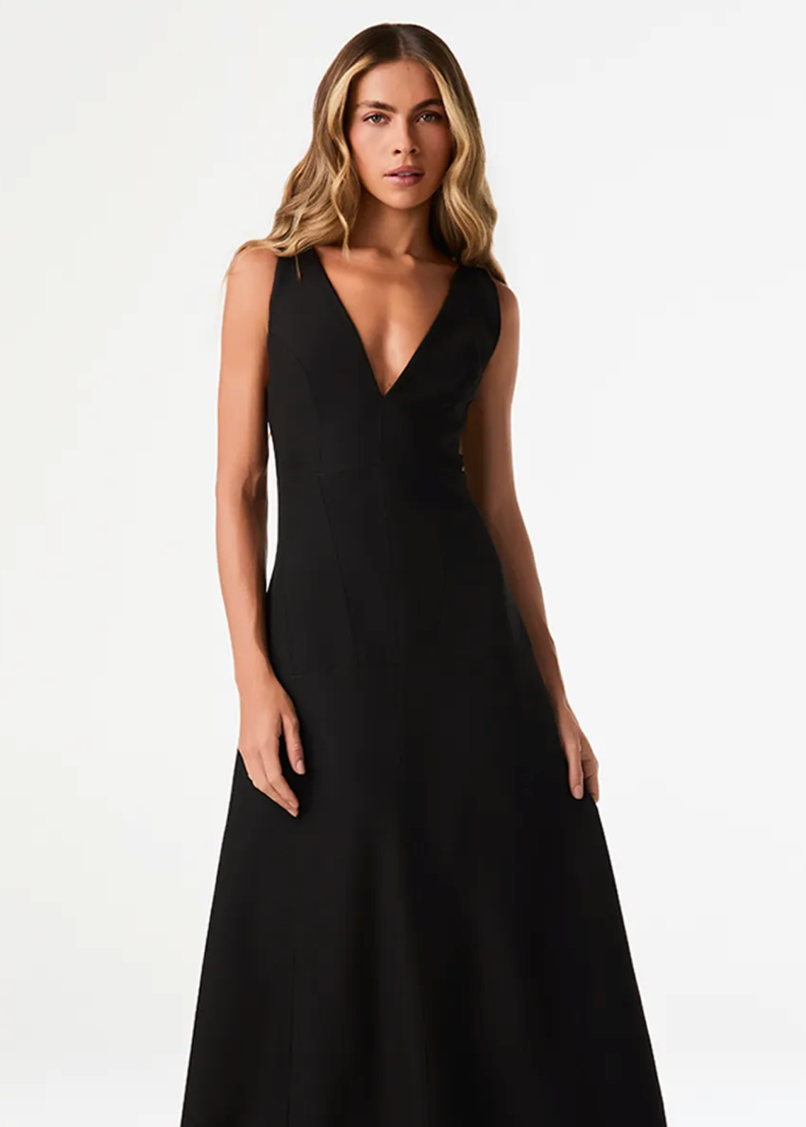 A woman modeling a sleeveless black A-line gown with a deep V-neckline, photographed against a clean white studio background.