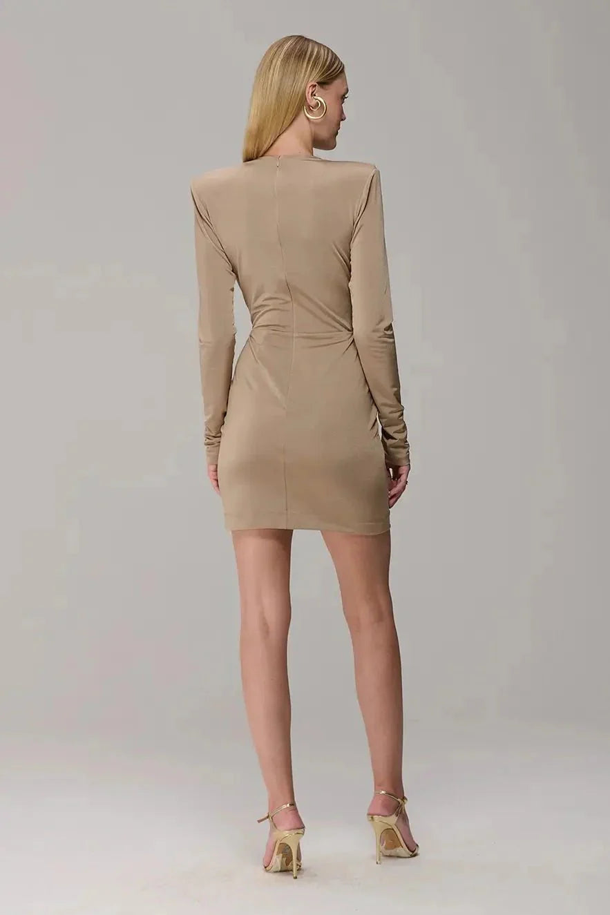 A woman viewed from the back wearing a fitted beige long-sleeve mini dress with structured shoulders and a ruched waist, paired with metallic gold high-heeled sandals against a light gray studio background.