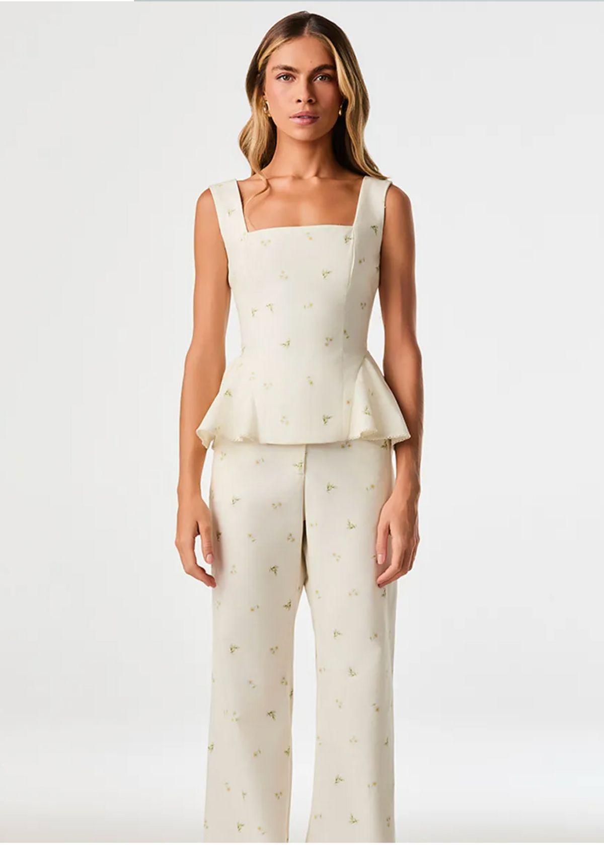 A model stands against a plain light gray studio background wearing a matching sleeveless peplum top and wide-leg pants set in warm off-white with a small scattered gold floral print.