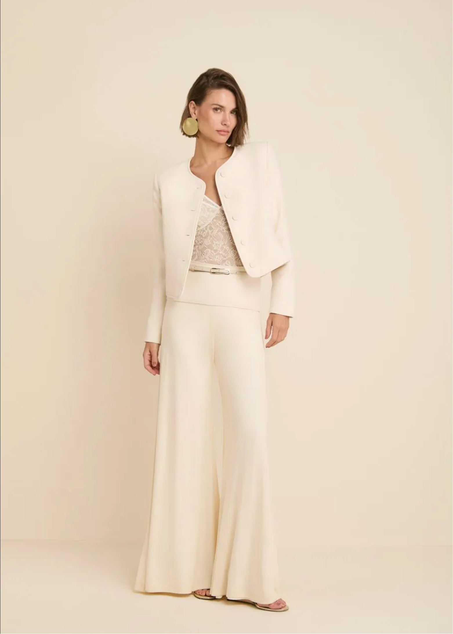 A woman models a cream-colored two-piece suit consisting of a cropped jacket and wide-leg trousers, layered over a lace top, against a solid cream background.