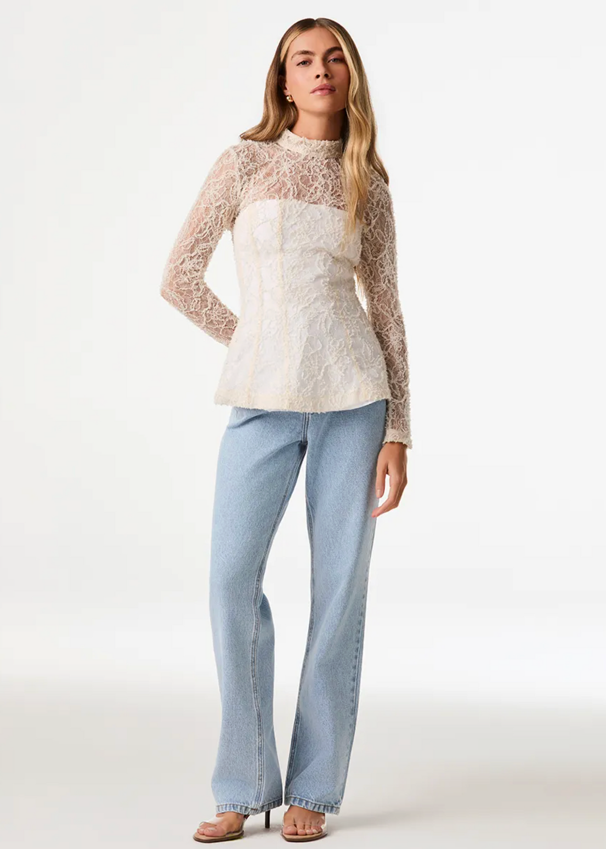 A woman modeling a cream lace long-sleeve top with a mock neck and light-wash blue jeans against a plain white studio background.