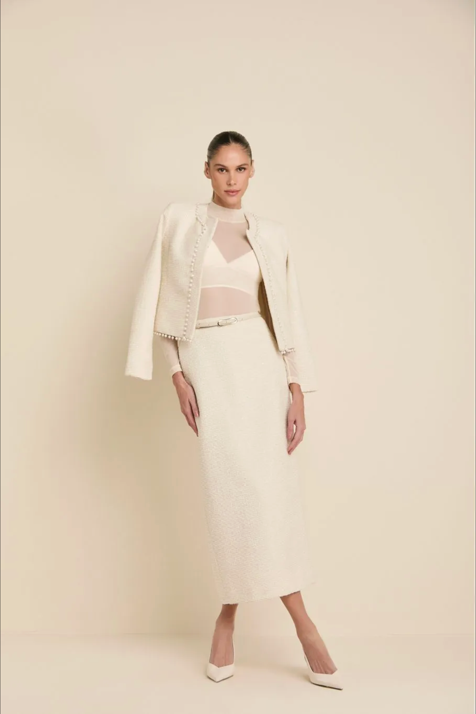 A model stands against a warm beige studio backdrop wearing a coordinated cream outfit: a cropped jacket with decorative trim, a matching midi skirt, a sheer underwire bra, and white pointed heels.