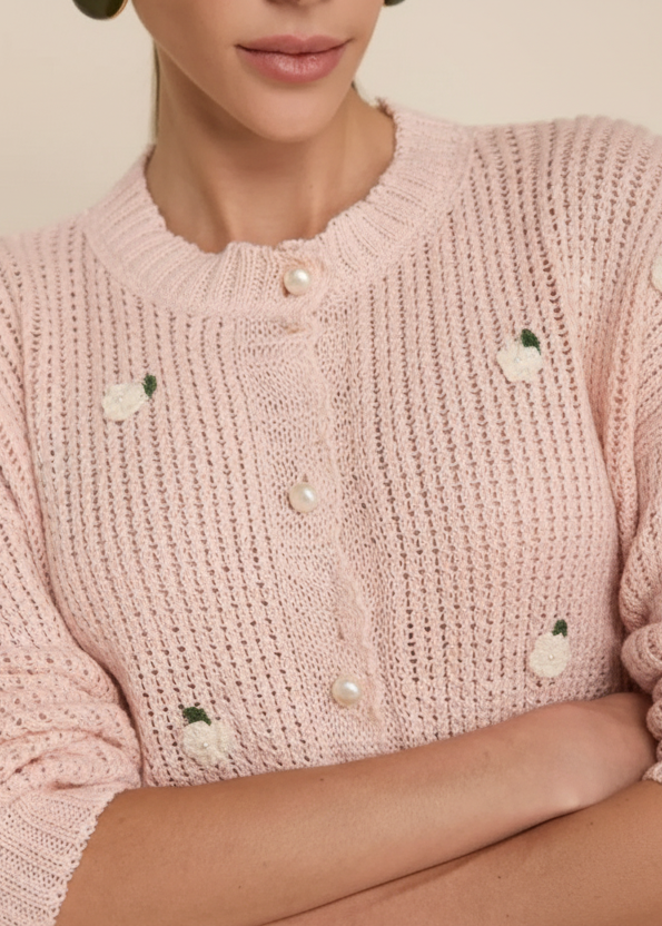 A close-up of a woman wearing a cream-colored knit cardigan sweater with small embroidered green leaves and white flower details, styled with pearl-like buttons.