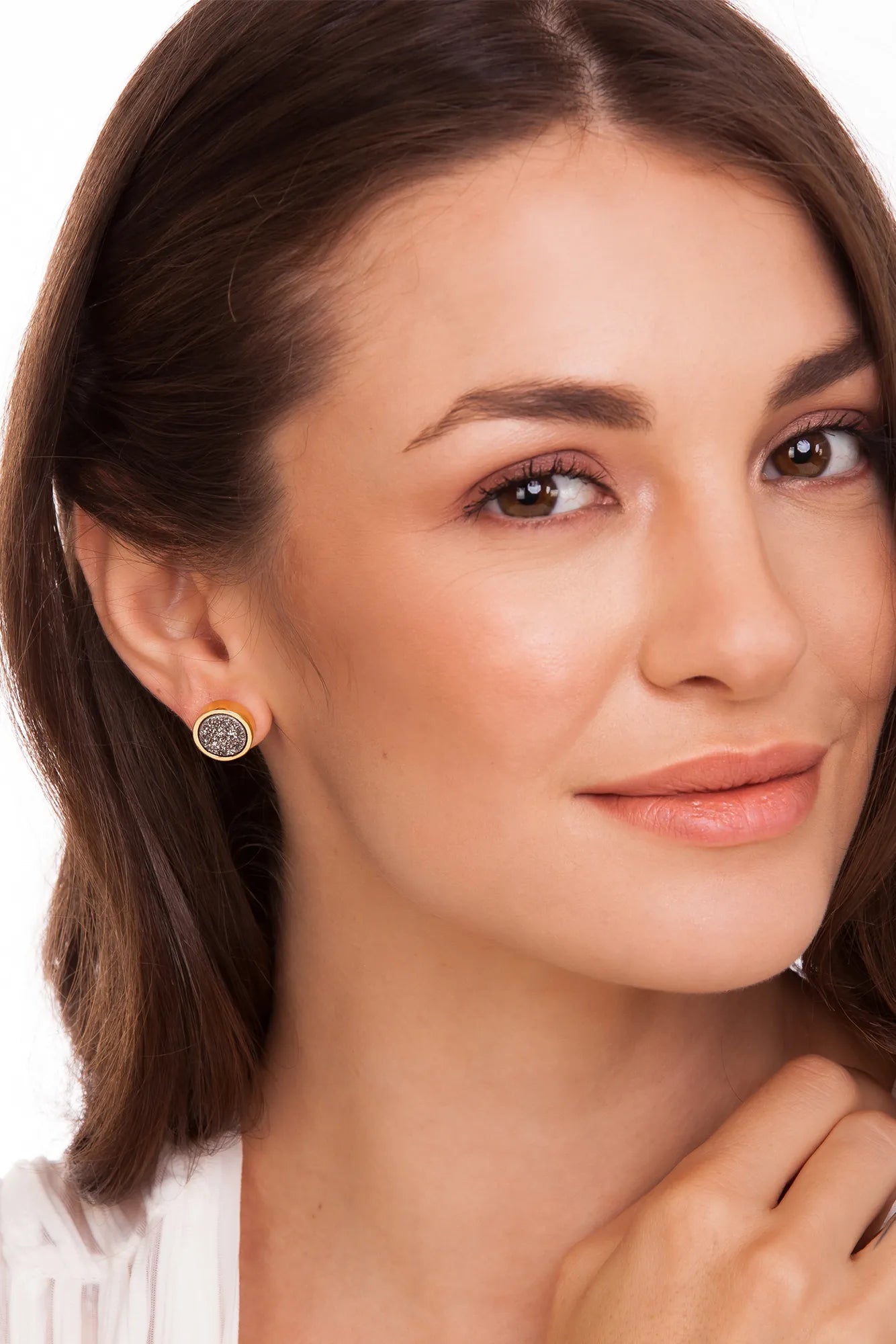 A close-up of a woman wearing a small gold stud earring with a circular, textured gray and white center, set against a clean white background.