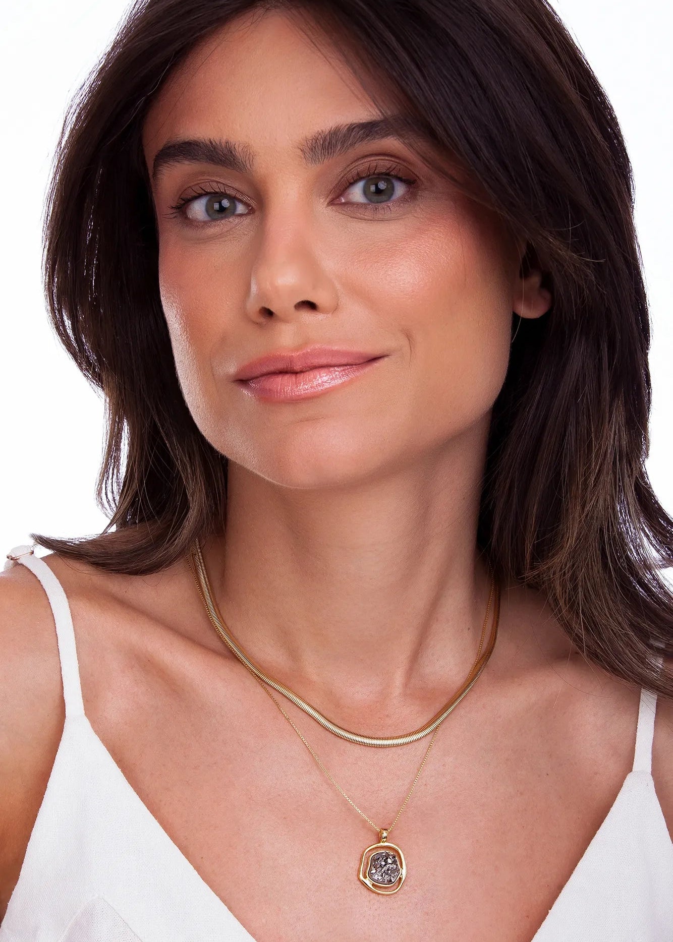 A close-up of a woman wearing a double-layered gold necklace, featuring a delicate chain and a larger chain with a small gold pendant containing clear crystals, set against a plain white background.