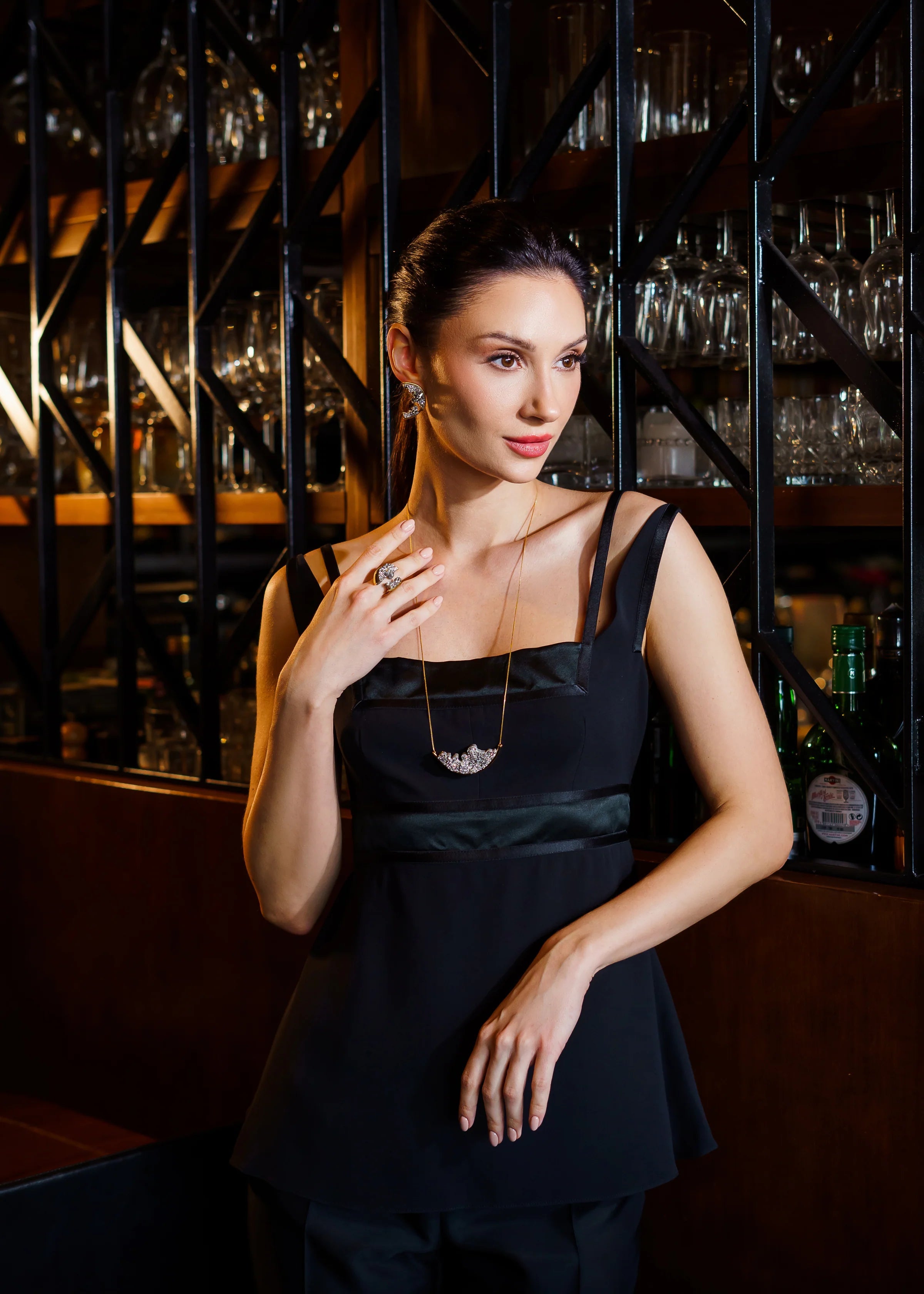 Woman in a black dress standing in a bar setting with shelves of glasses and bottles.