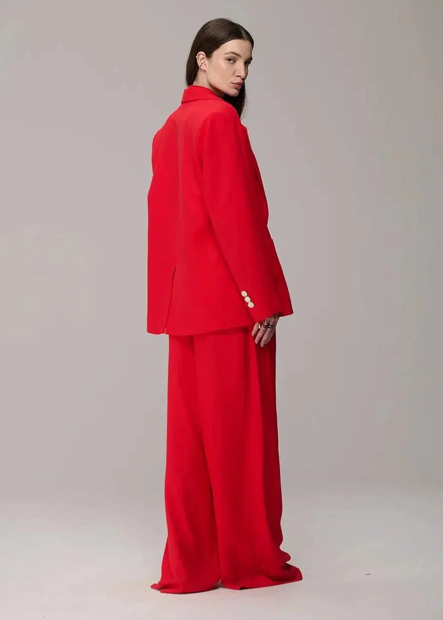 A model is shown from the back wearing a bright red two-piece suit consisting of a structured blazer with three decorative buttons on the sleeve and matching wide-leg trousers, set against a plain light gray studio background.