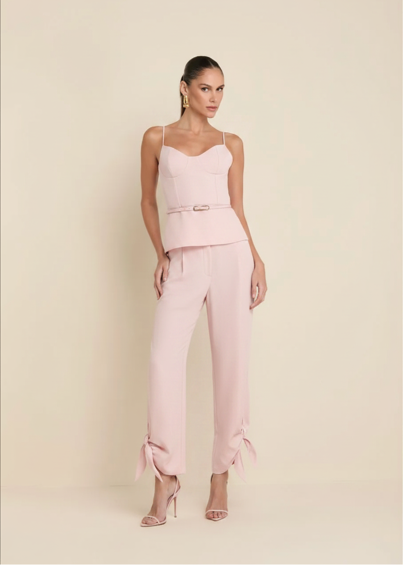 A model stands against a solid beige studio background wearing pink sleeveless jumpsuit with a structured corset-style bodice, a matching belt, and wide-leg cropped pants with tie details at the ankles.