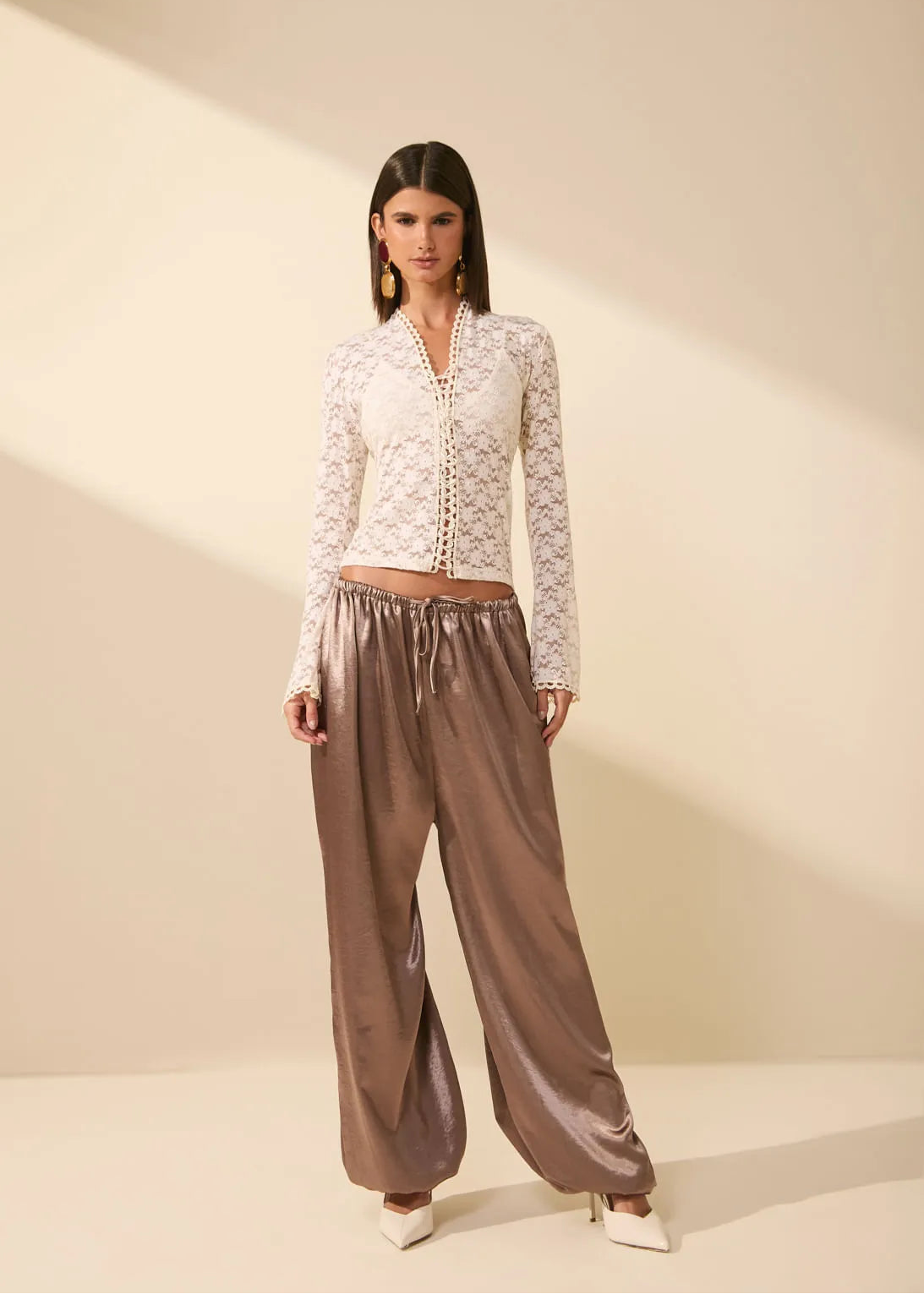 A model stands in a studio wearing a white lace long-sleeve crop top and shiny taupe-brown wide-leg pants with white pointed shoes.