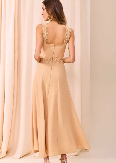Woman wearing a beige evening gown with lace detailing in front of beige curtains.