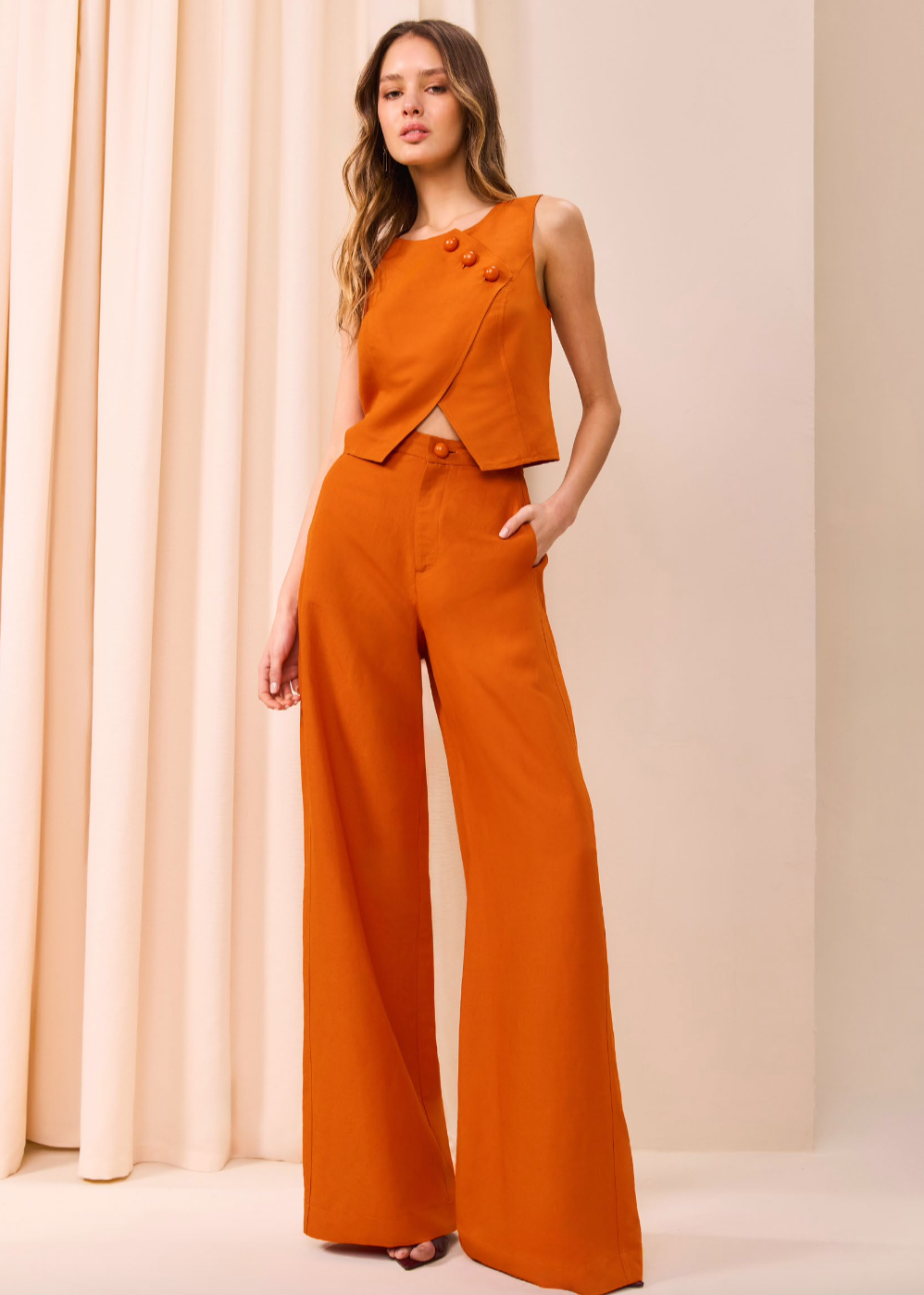 Woman wearing an orange outfit with a matching top and wide-leg pants against a beige curtain background.