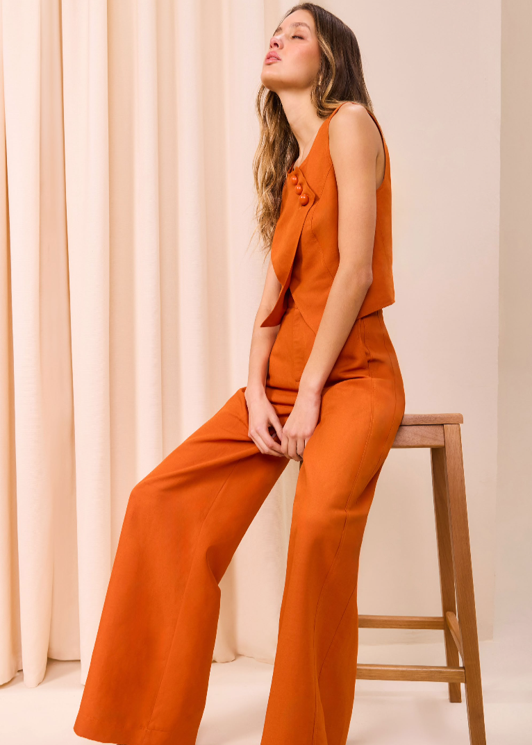 Woman in an orange jumpsuit sitting on a wooden stool against a beige curtain.
