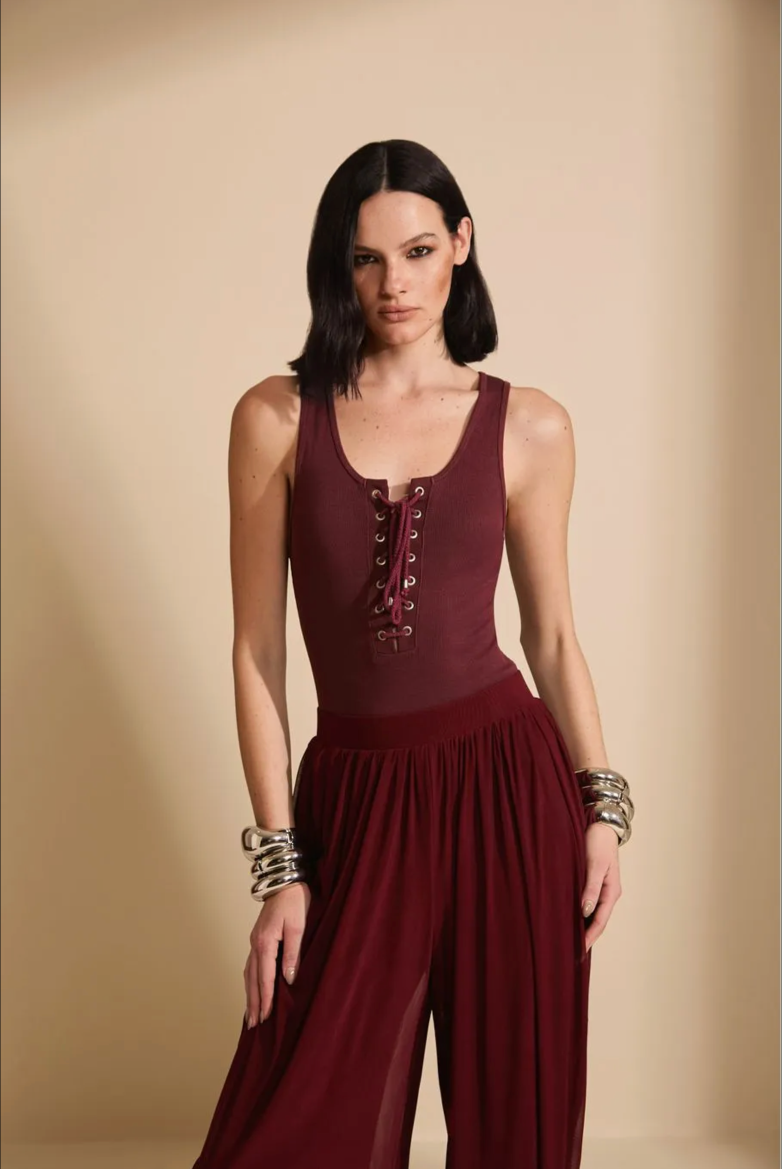 A woman modeling a sleeveless deep burgundy jumpsuit with lace-up detailing on the bodice and a flowing skirt, posed against a solid beige studio backdrop.