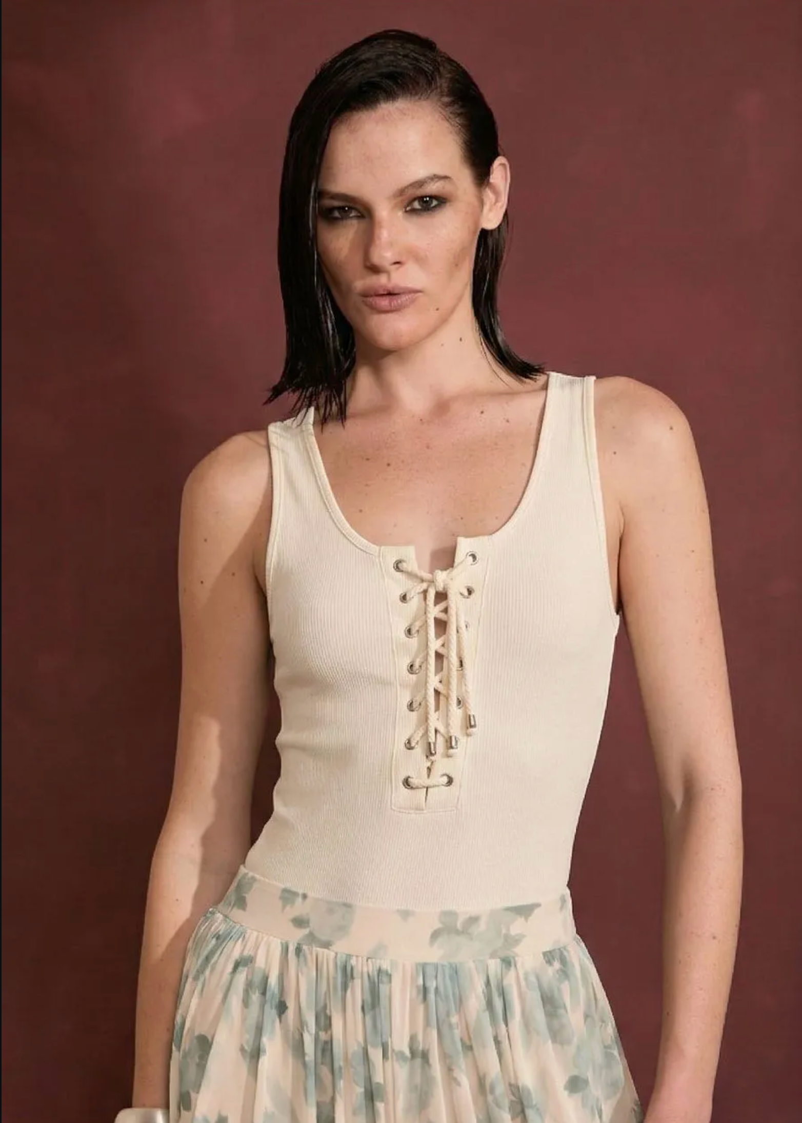 A model wearing a cream-colored ribbed knit lace-up tank top and a light blue-green floral print skirt, standing against a solid dark reddish-brown background.