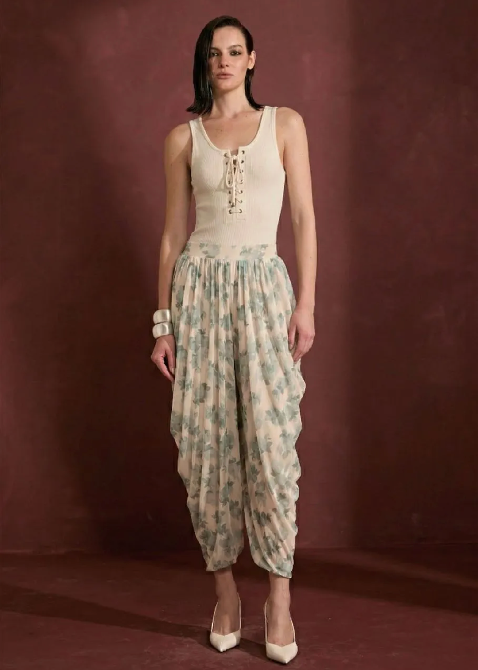 A model stands against a textured dark reddish-brown studio backdrop wearing a cream-colored lace-up tank top and matching cream harem pants with a light blue-green floral print. She is wearing cream pointed-toe heels and a wide cream cuff bracelet.