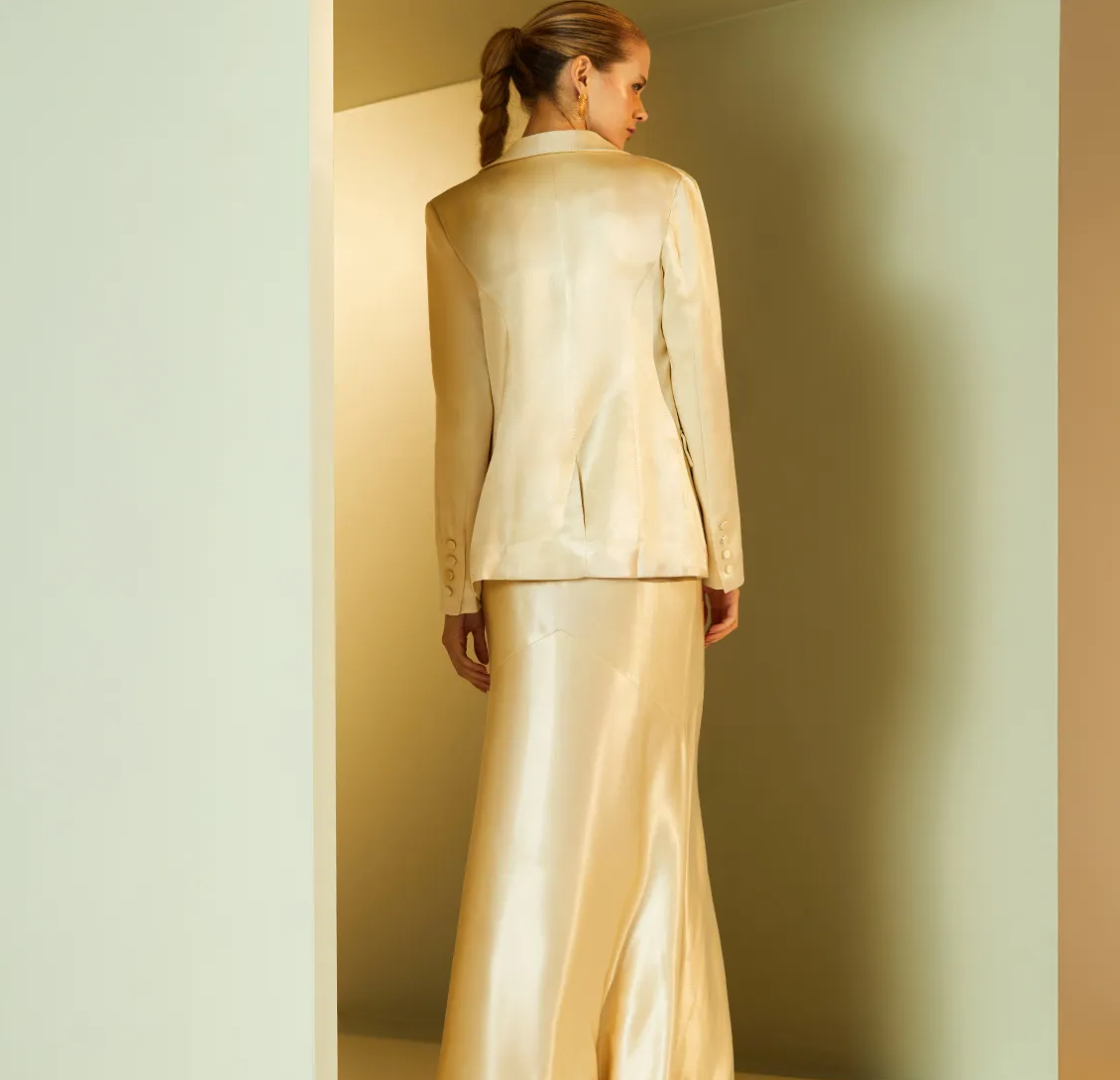 Woman wearing a light-colored suit standing against a plain wall.