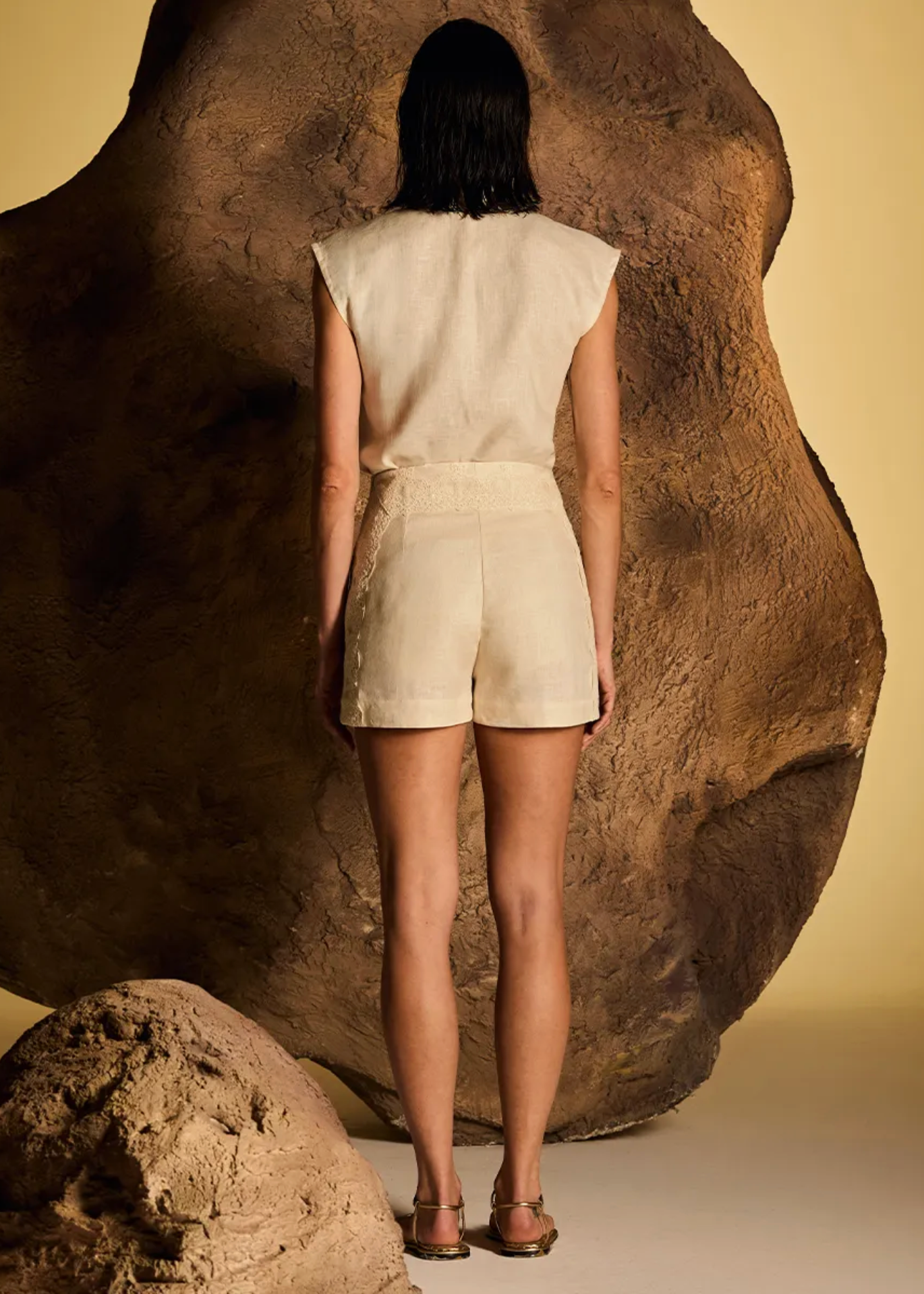 A model stands with her back to the camera wearing a cream-colored sleeveless top and matching shorts, positioned in front of a large textured brown rock prop against a warm beige background.