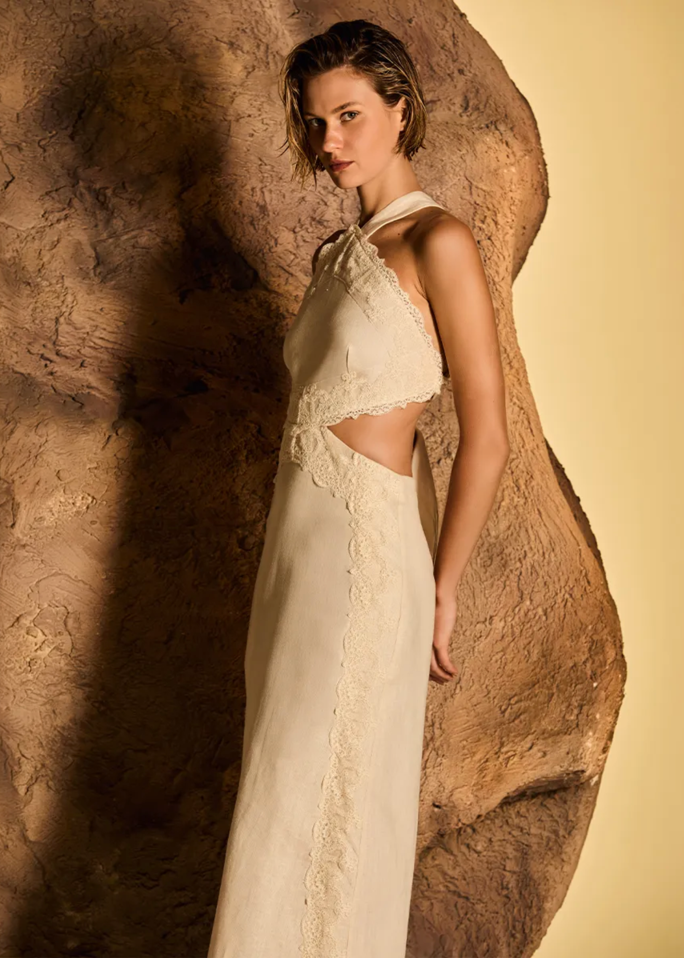 A model wearing a cream-colored halter-neck dress with lace details and side cutouts, posing in front of a large textured rock against a warm beige background.