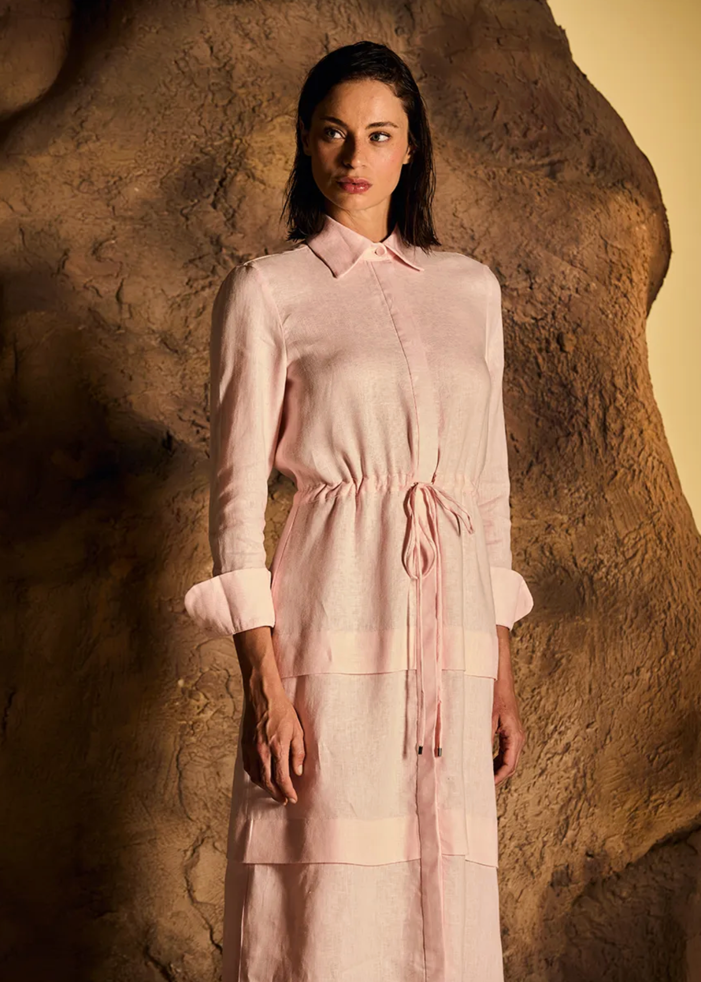 A woman stands in front of a textured rock formation wearing a light pink long-sleeve collared dress with a matching tie belt. The background is a warm, earthy brown rock with a yellow light on the right side.